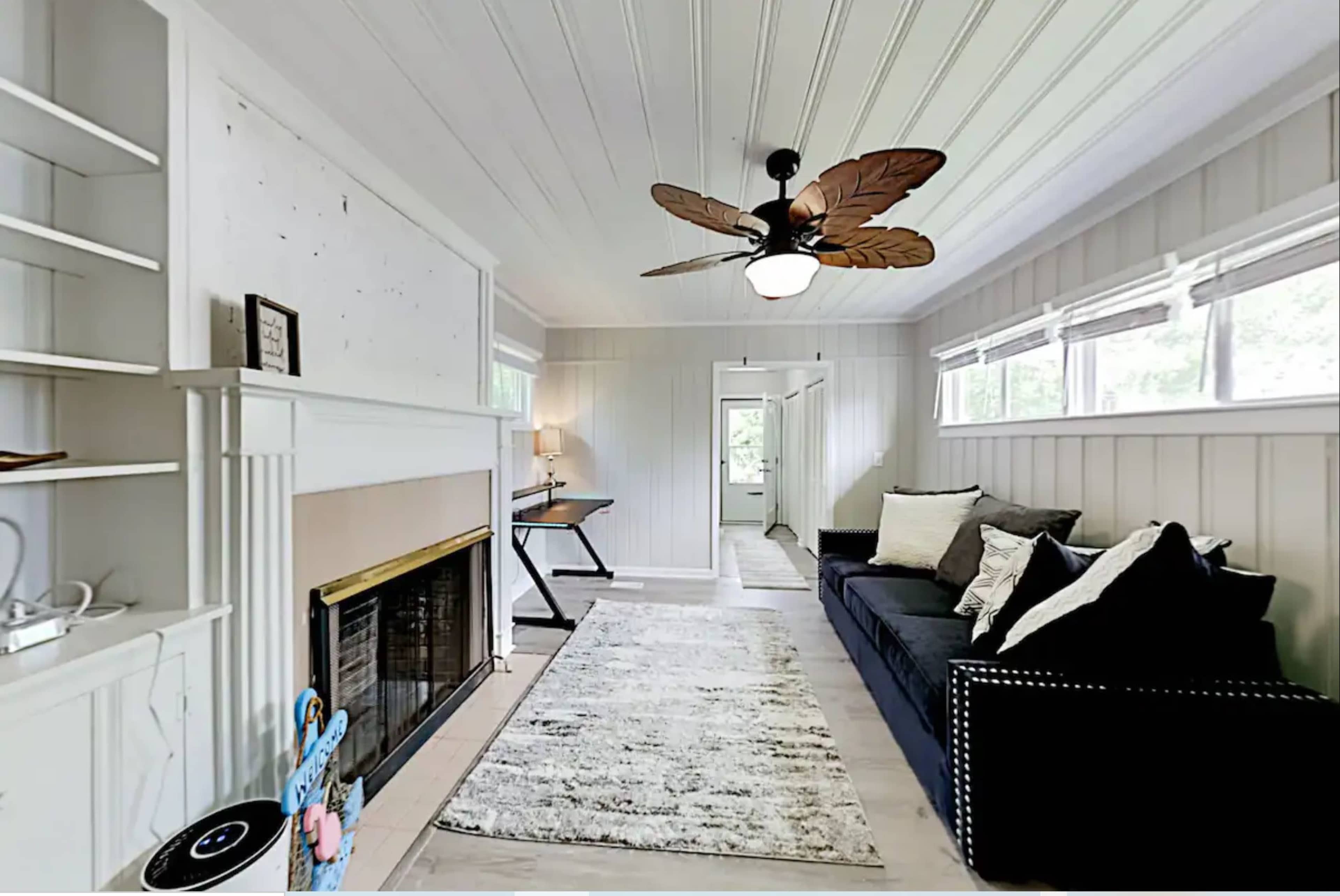 The image shows a well-lit living room featuring a black sofa, a white fireplace, and a large rug, with a ceiling fan overhead and a desk visible in the background.