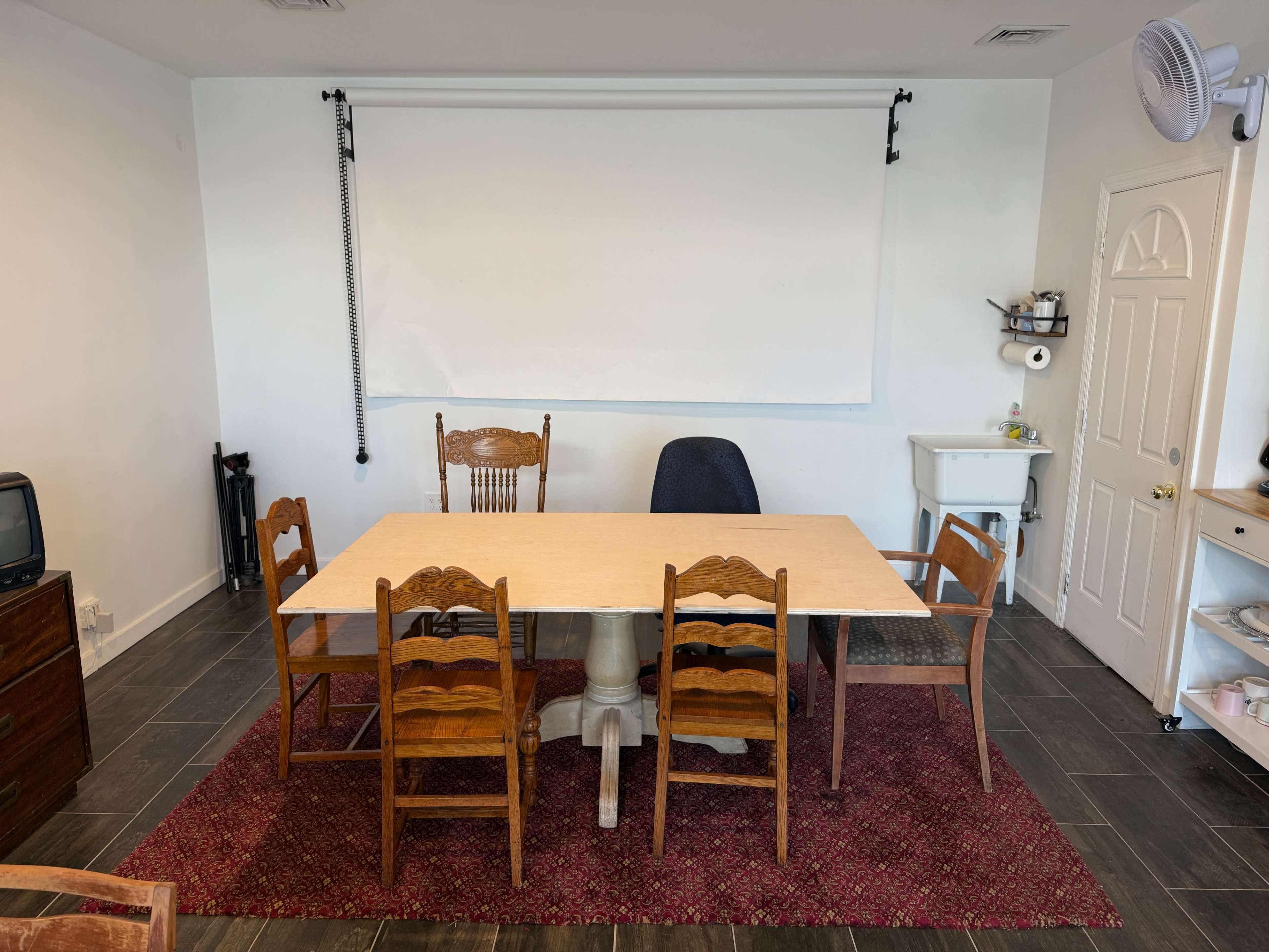 A large wooden table with six chairs is centered in a room that features a whiteboard and a small cabinet, with a vintage television set in the corner.