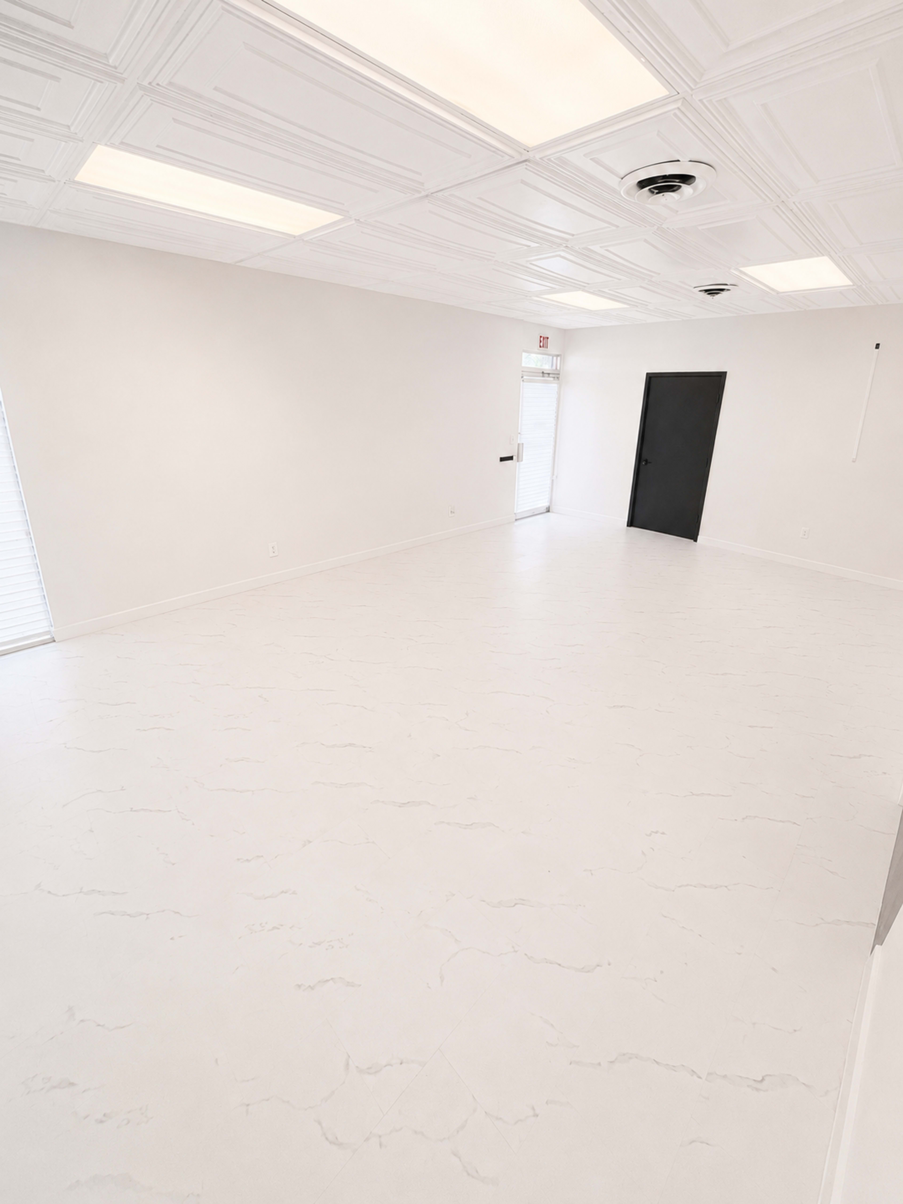 The image shows a spacious, empty room with a white floor and walls, featuring a black door and several ceiling lights.