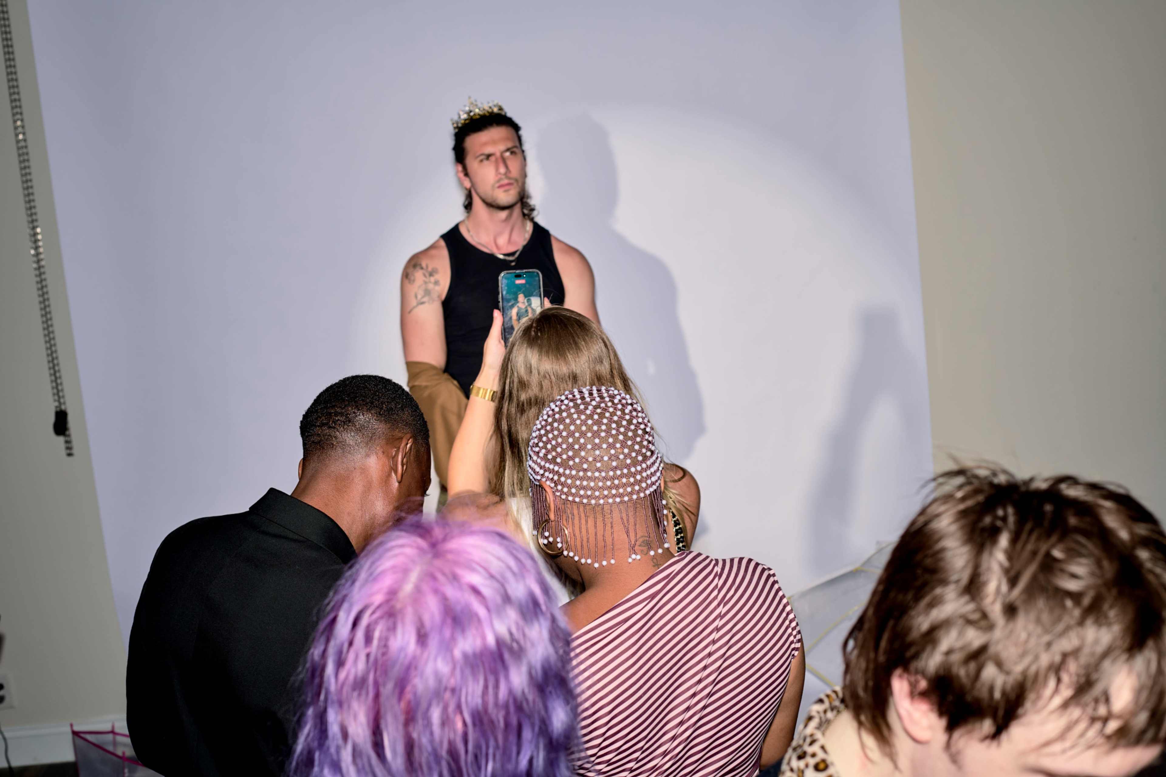 A group of people is focused on a man in a tank top wearing a crown while he stands against a plain backdrop.