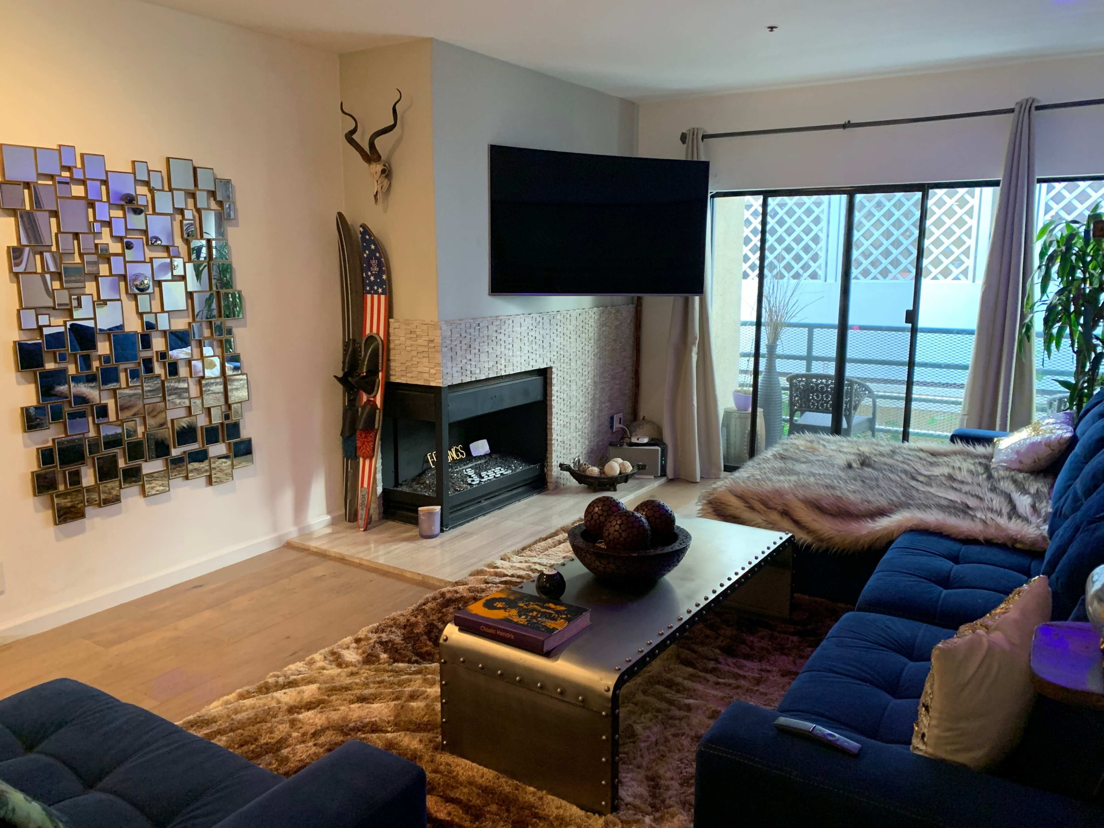 The living room features a modern design with a blue sectional sofa, a coffee table, a fireplace, and a large mirror wall alongside a balcony door.