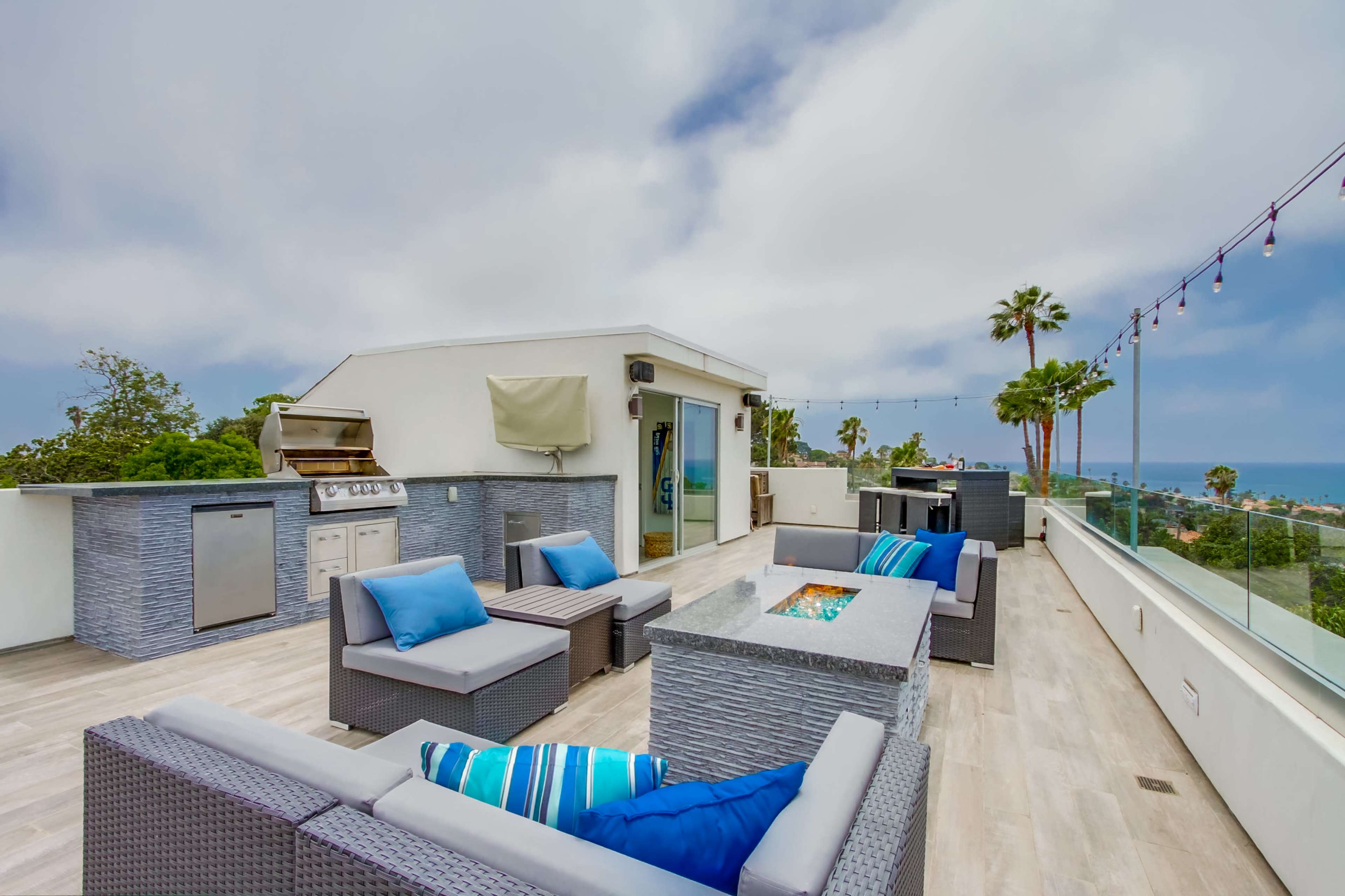 A rooftop terrace with modern outdoor furniture, a barbecue grill, and a view of the ocean under a cloudy sky.
