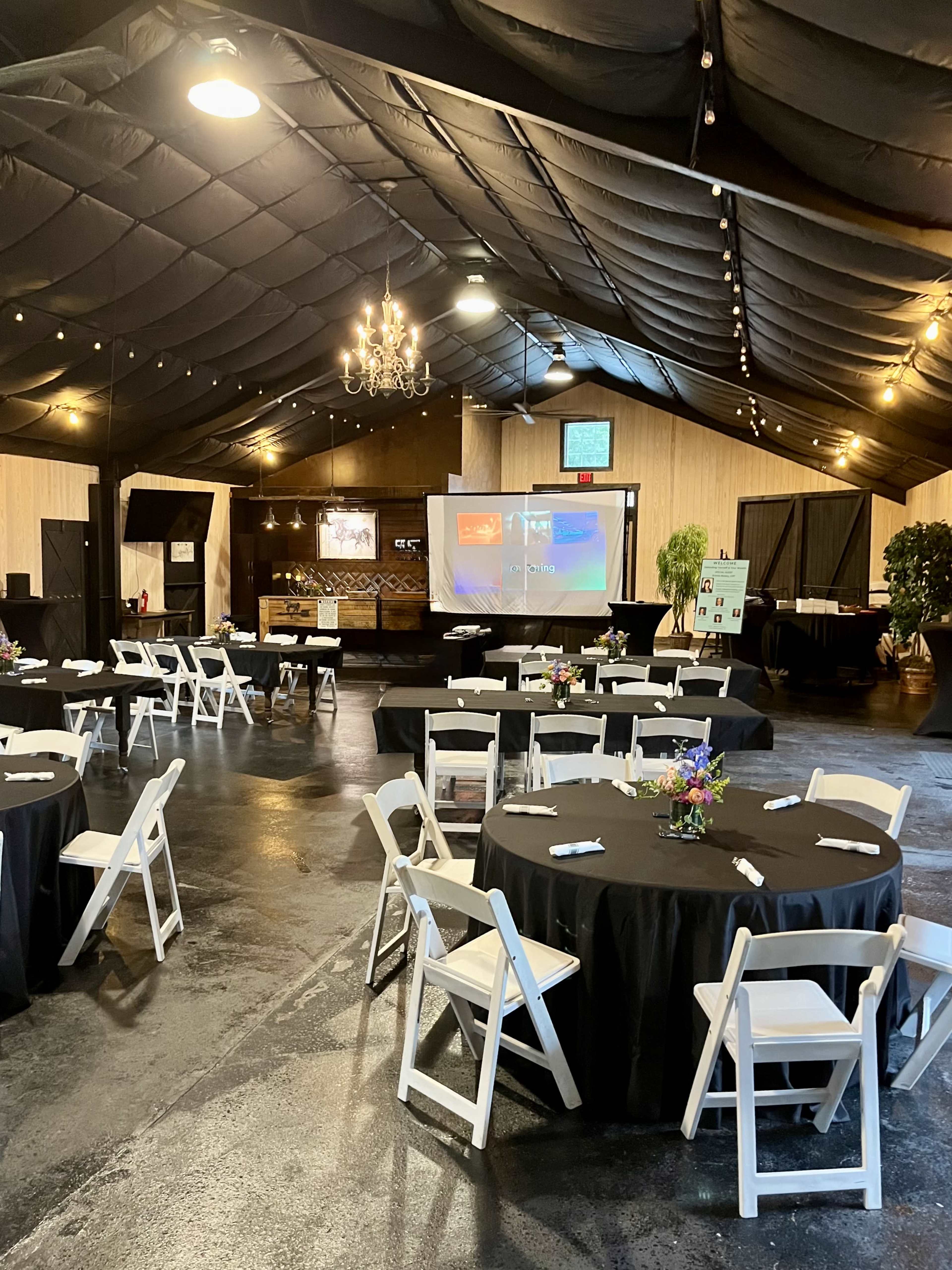 The image shows a spacious indoor event venue with round tables covered in black tablecloths, a large screen for presentations, and decorative lighting hanging from a black ceiling.