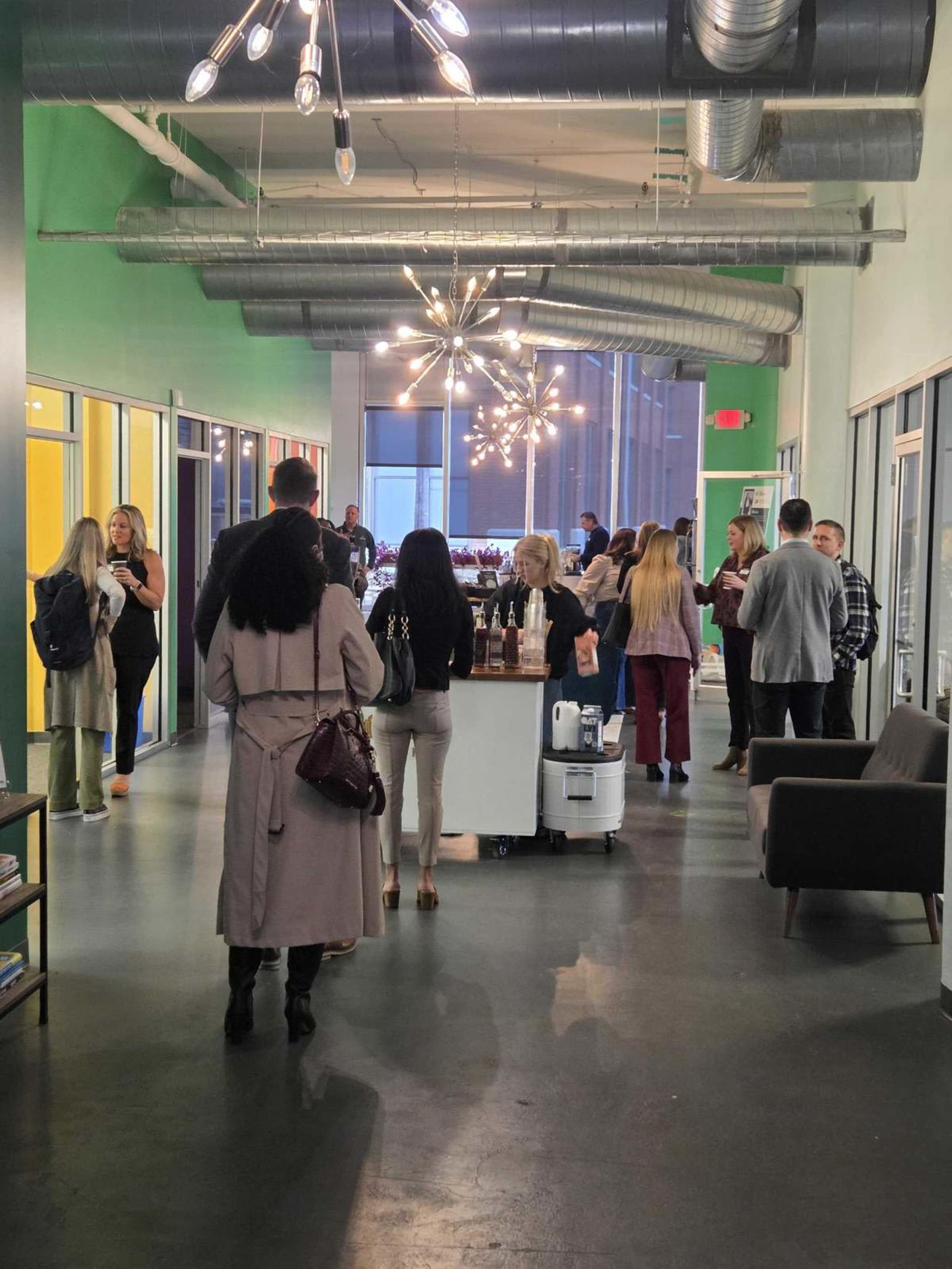 People are mingling at a networking event in a brightly lit hallway with modern decor and refreshments displayed on a table.