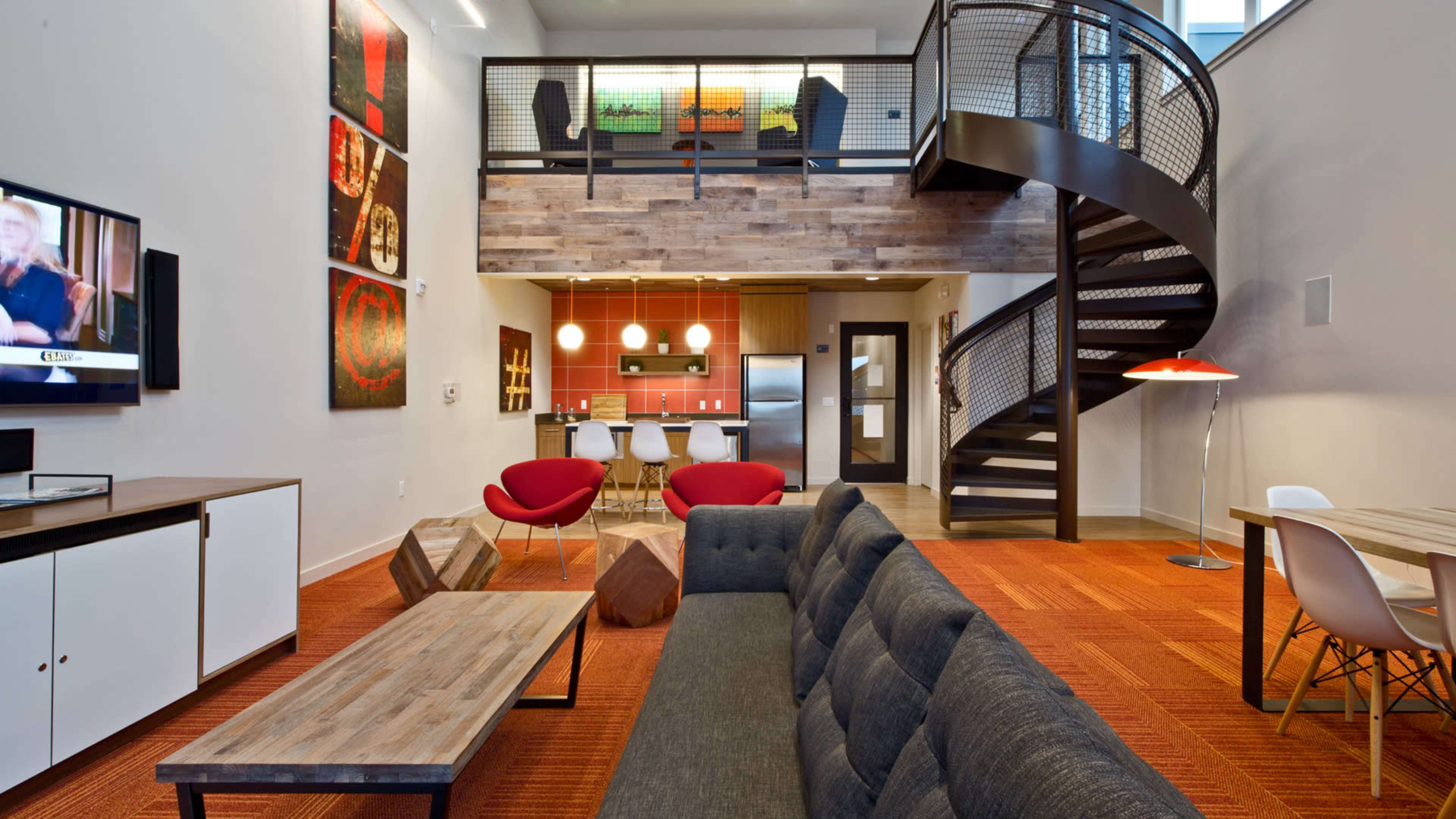 The image shows a modern living space featuring a grey sofa, a wooden coffee table, a staircase, and a kitchen area with bar stools and colorful wall art.