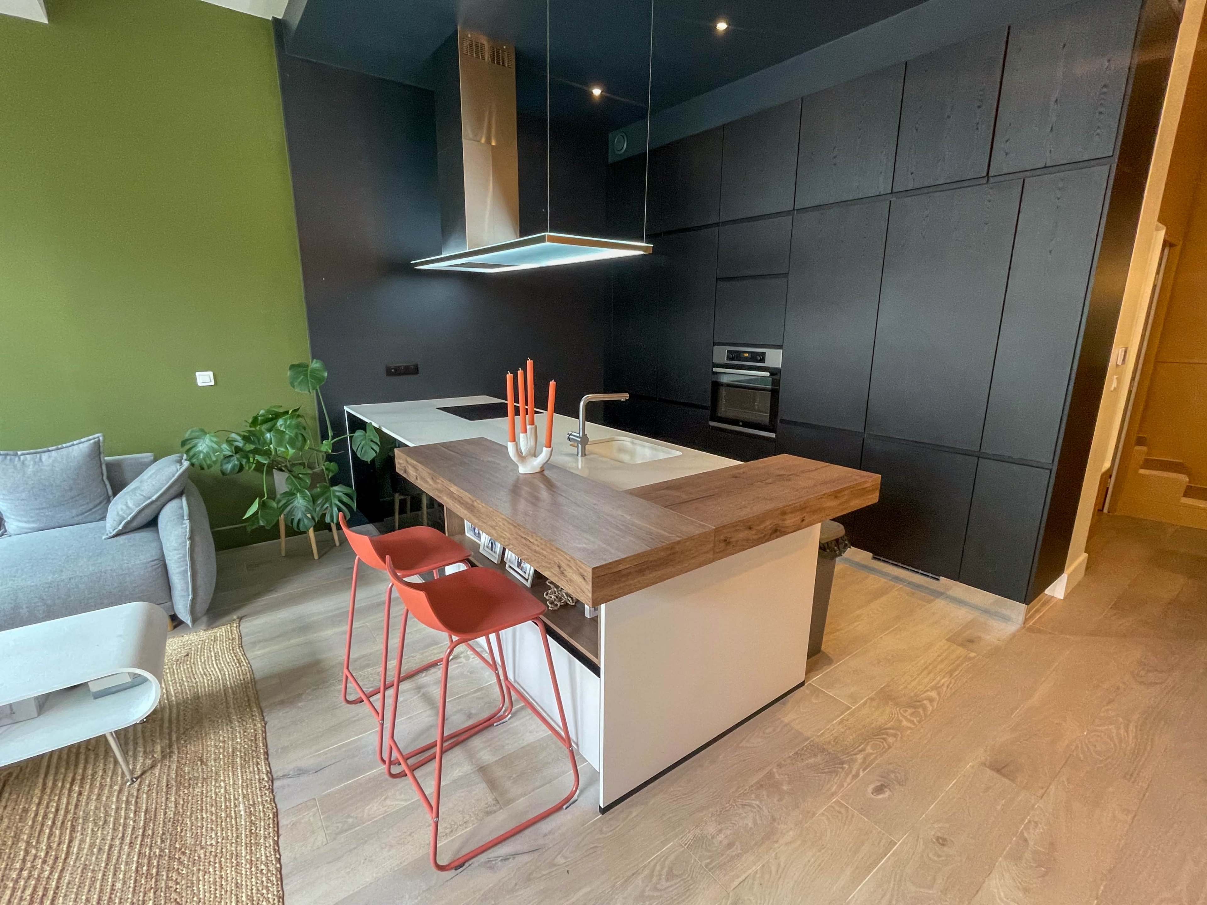 The image shows a modern kitchen featuring a central island with a wooden countertop, two red bar stools, and dark cabinetry, set against a green accent wall.