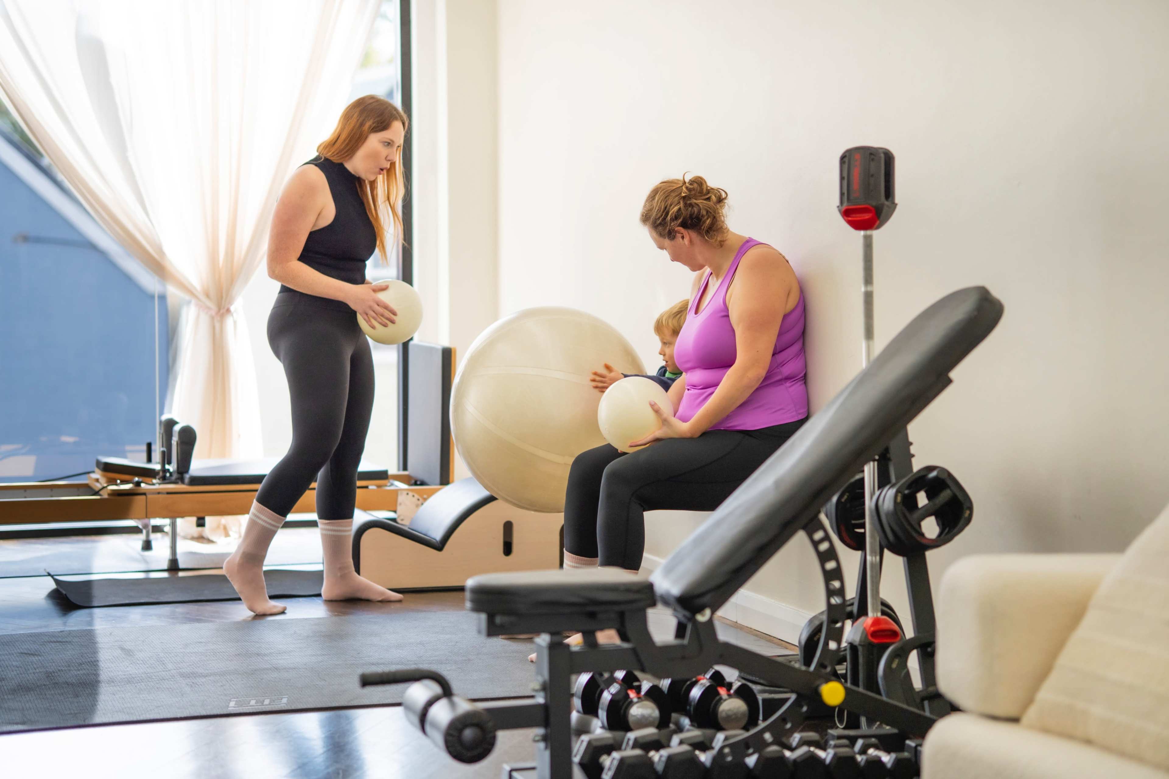 Two women are in a fitness studio, one holding a stability ball while the other sits on a bench with a child beside her.