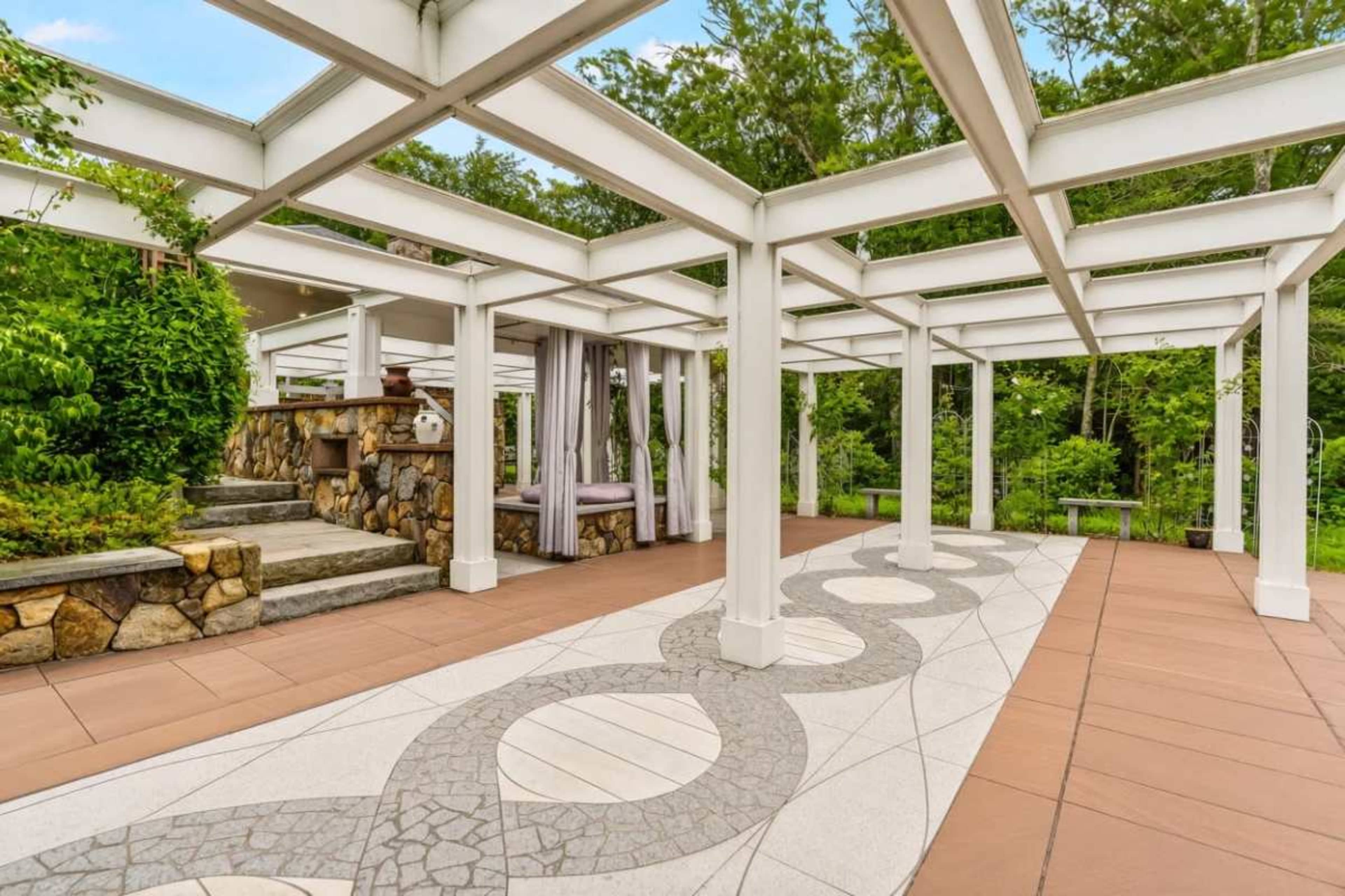 The image shows a spacious outdoor patio area surrounded by greenery, featuring a stone wall, a seating area with curtains, and a decorative tiled pathway.