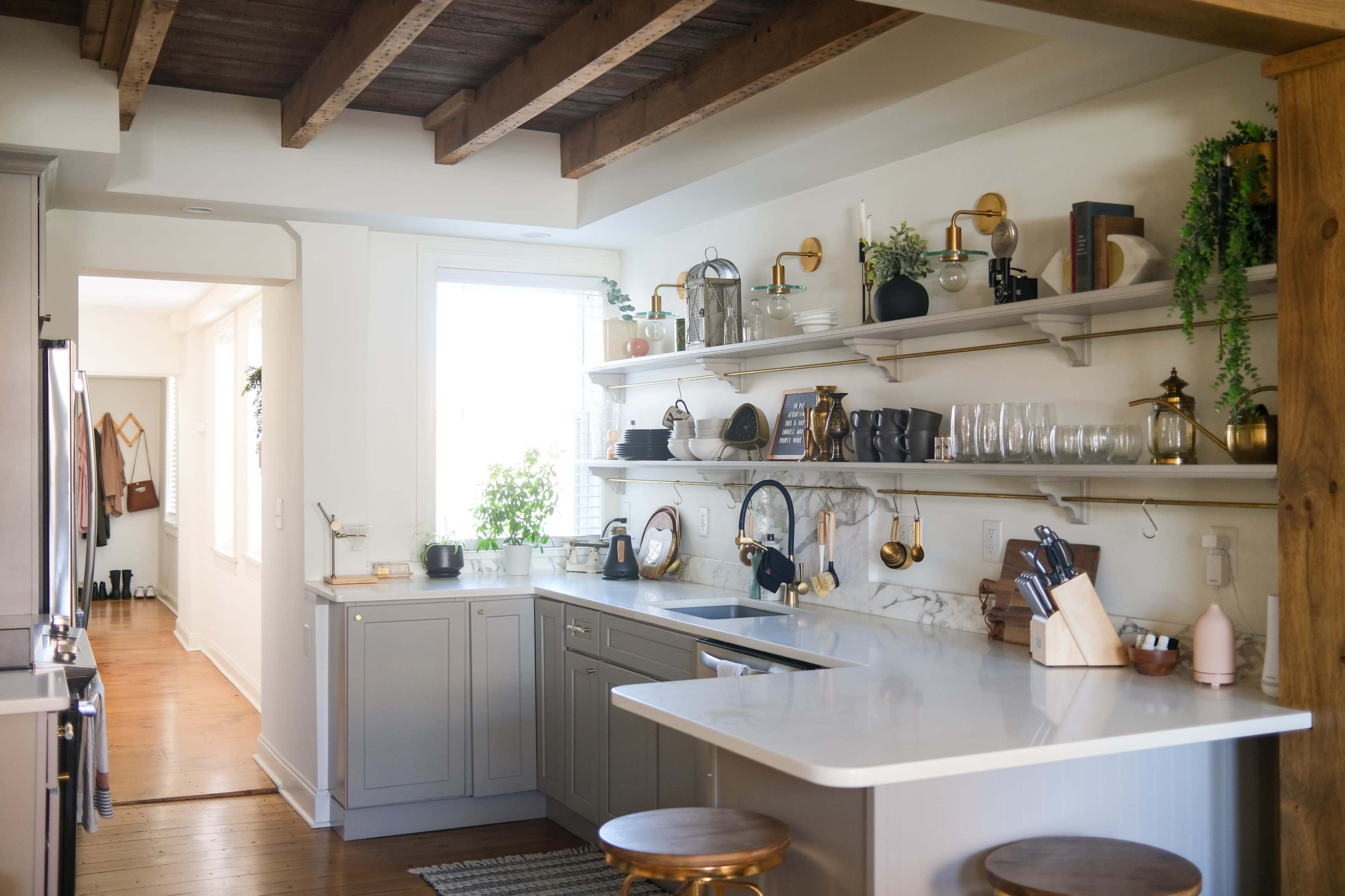 A modern kitchen with gray cabinets, a white countertop, open shelving displaying kitchenware, and wooden beams overhead.