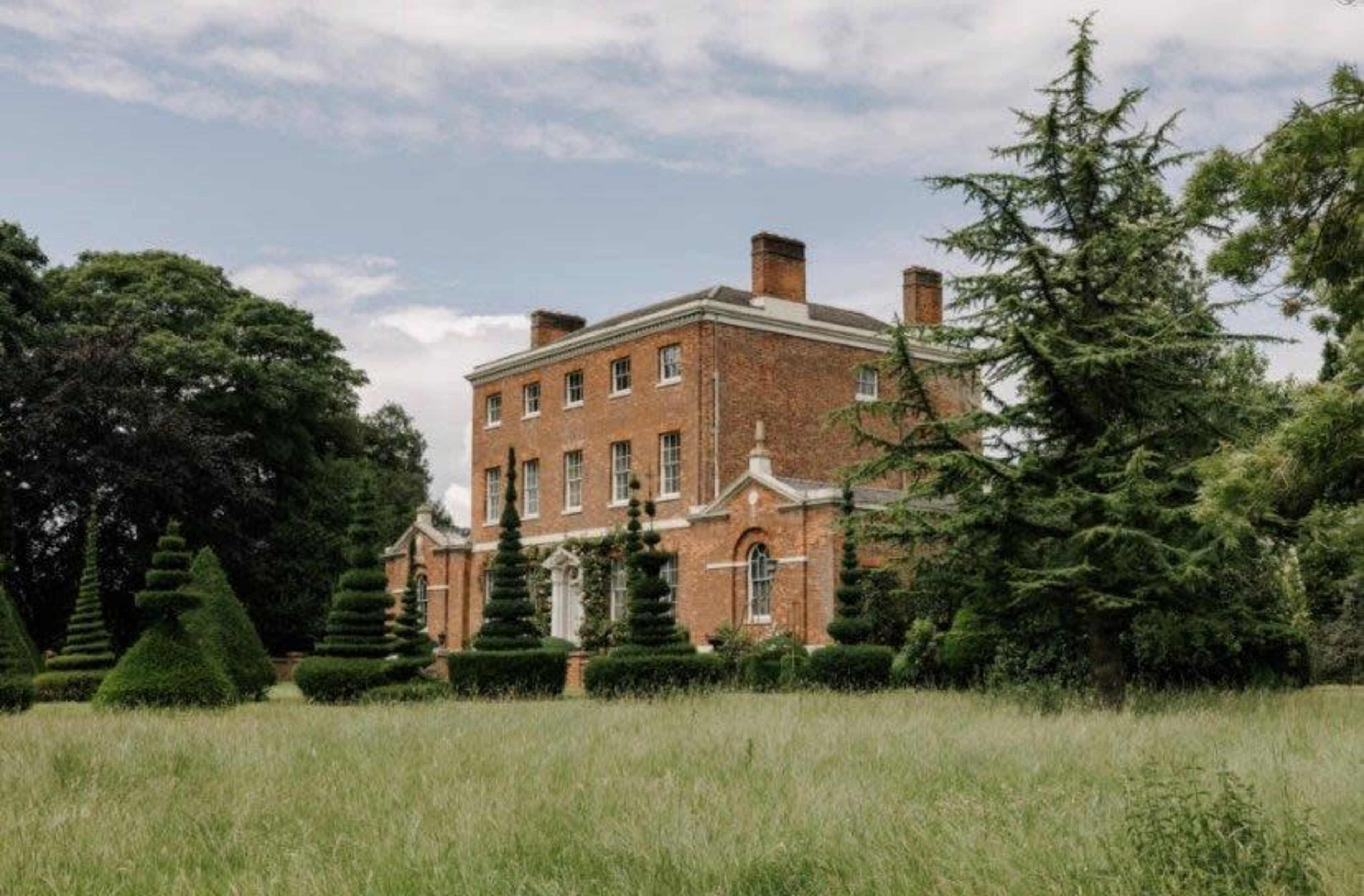 A large brick mansion with symmetrical windows is surrounded by manicured lawns and conical topiary trees.