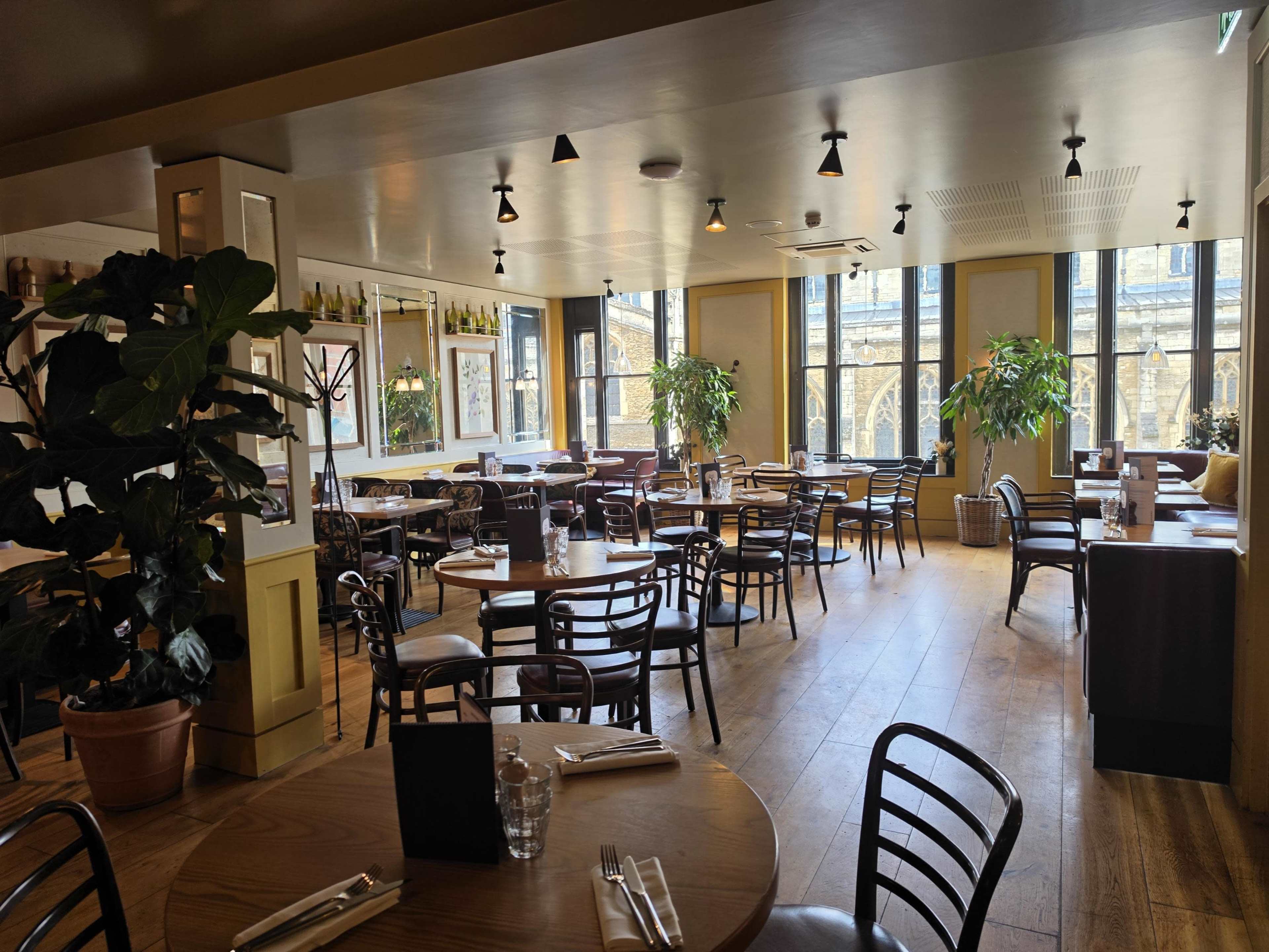 The image shows a spacious restaurant with wooden floors, several tables and chairs, and large windows letting in natural light, creating a bright atmosphere.