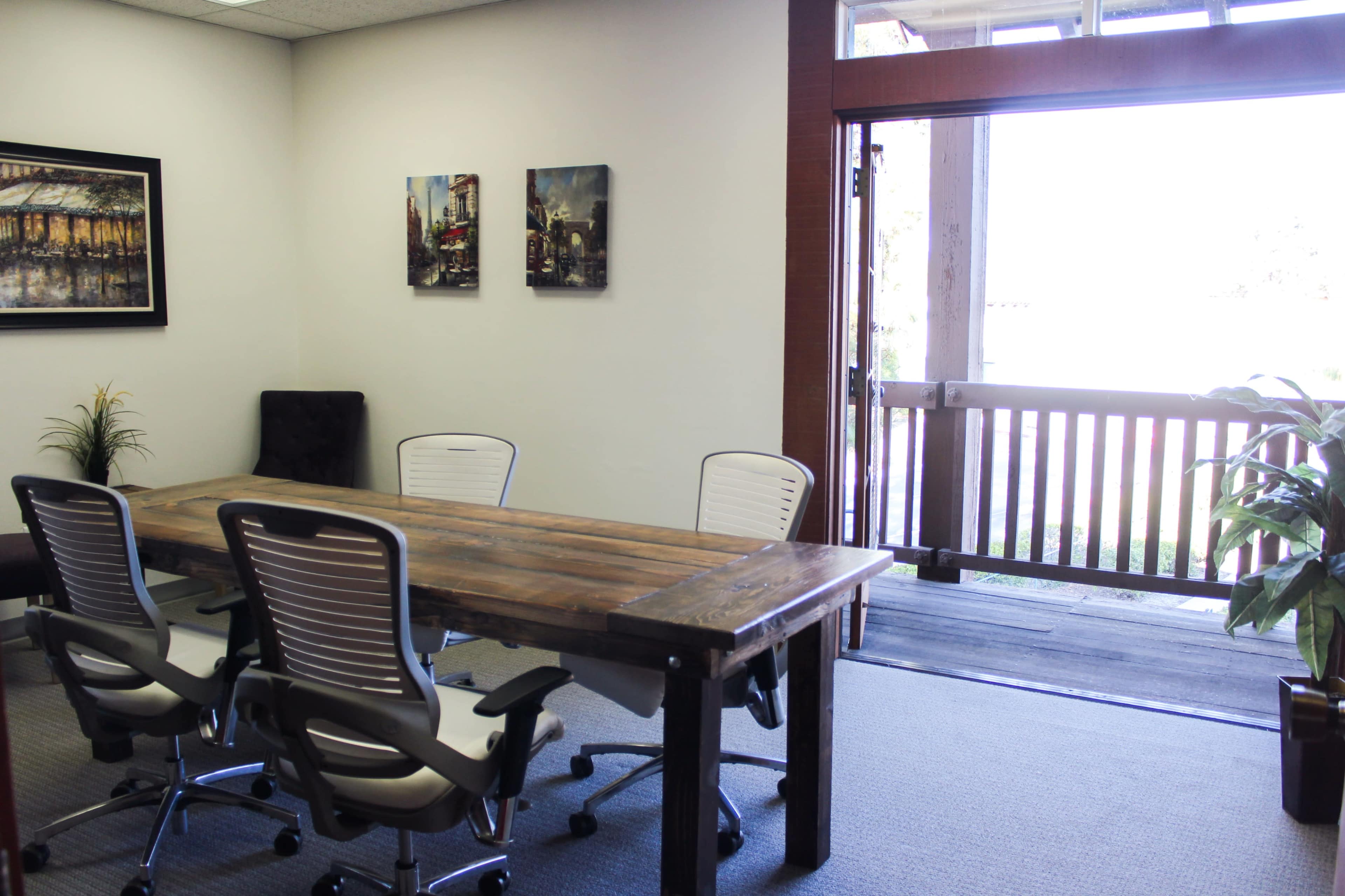 A conference room features a wooden table surrounded by white chairs, with a door leading to an outdoor area visible in the background.