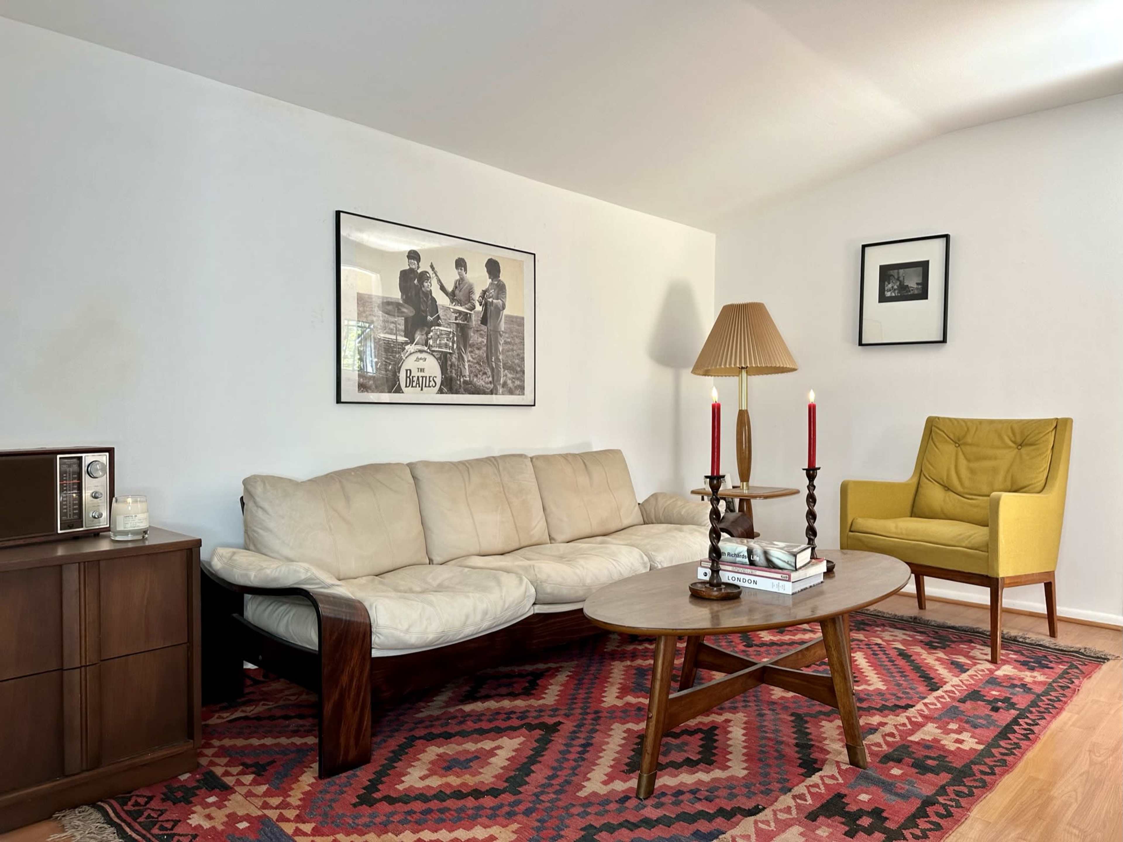 A living room with a beige couch, a wooden coffee table, a yellow armchair, and a decorative rug, accompanied by framed pictures on the walls.