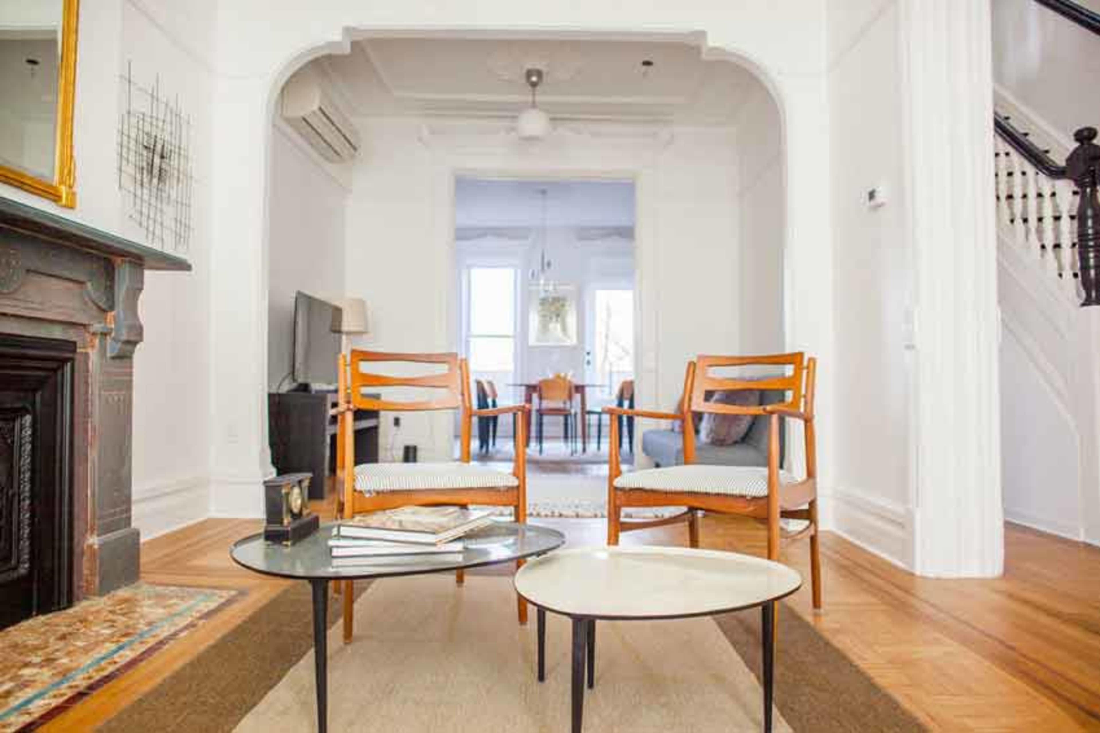 The image shows a spacious living room with two wooden chairs and two coffee tables, framed by a decorative archway and illuminated by natural light.