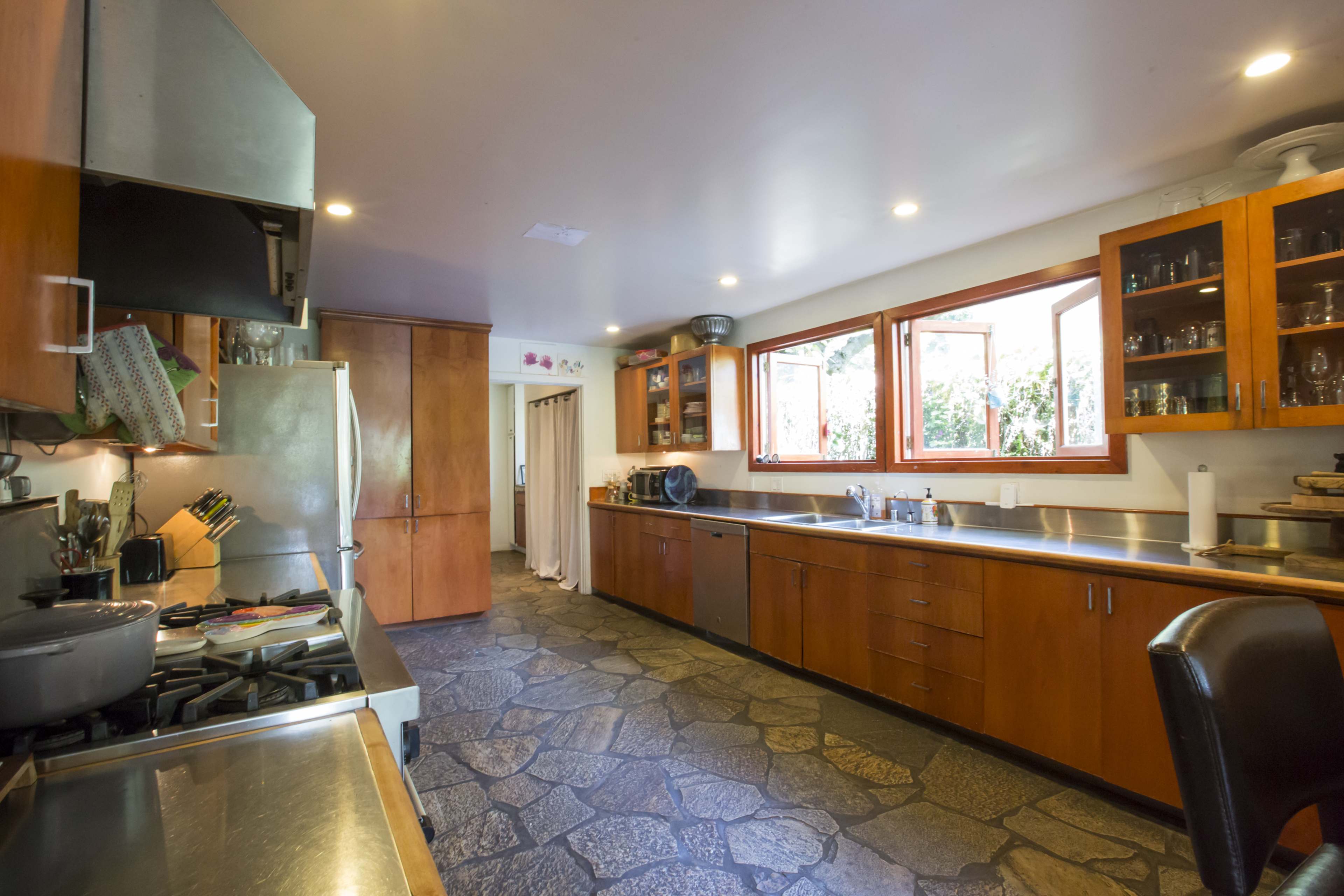 The kitchen features wooden cabinets, a stone floor, and large windows overlooking a garden area.