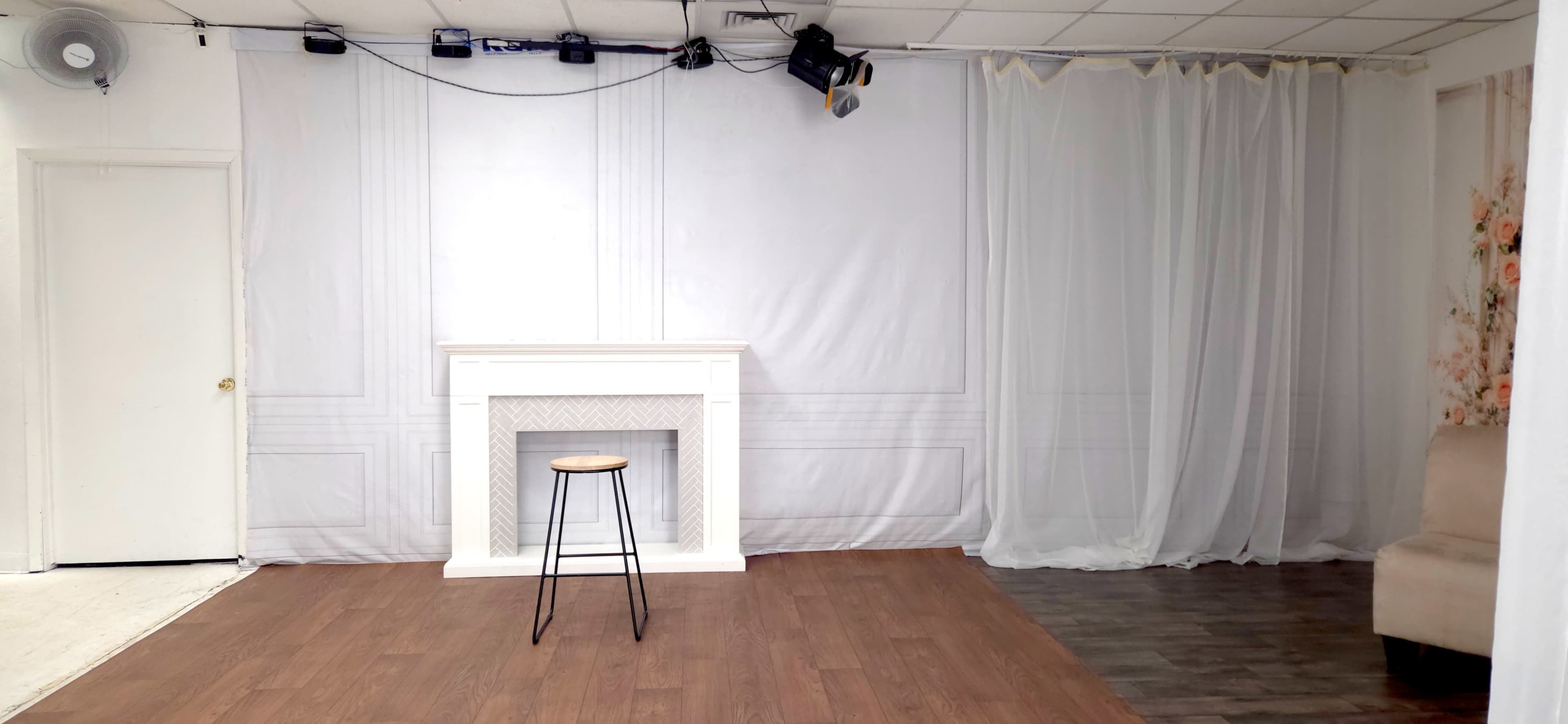 The image shows a simple room featuring a white wall with a faux fireplace and a single wooden stool in the center, accompanied by a light curtain and wooden flooring.