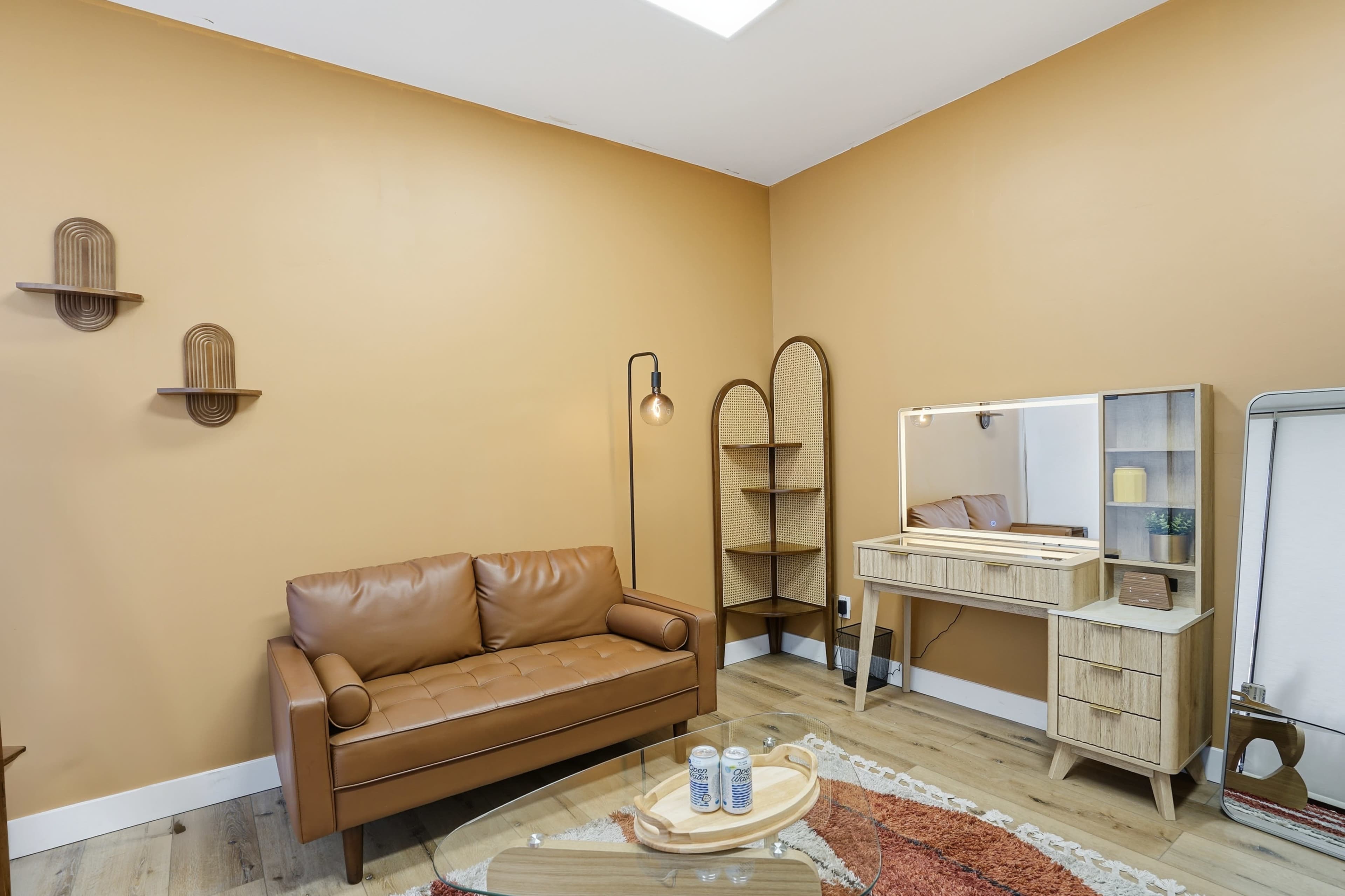 The image shows a cozy room with a brown leather sofa, a wooden shelf, a vanity with a mirror, and a warm orange wall.