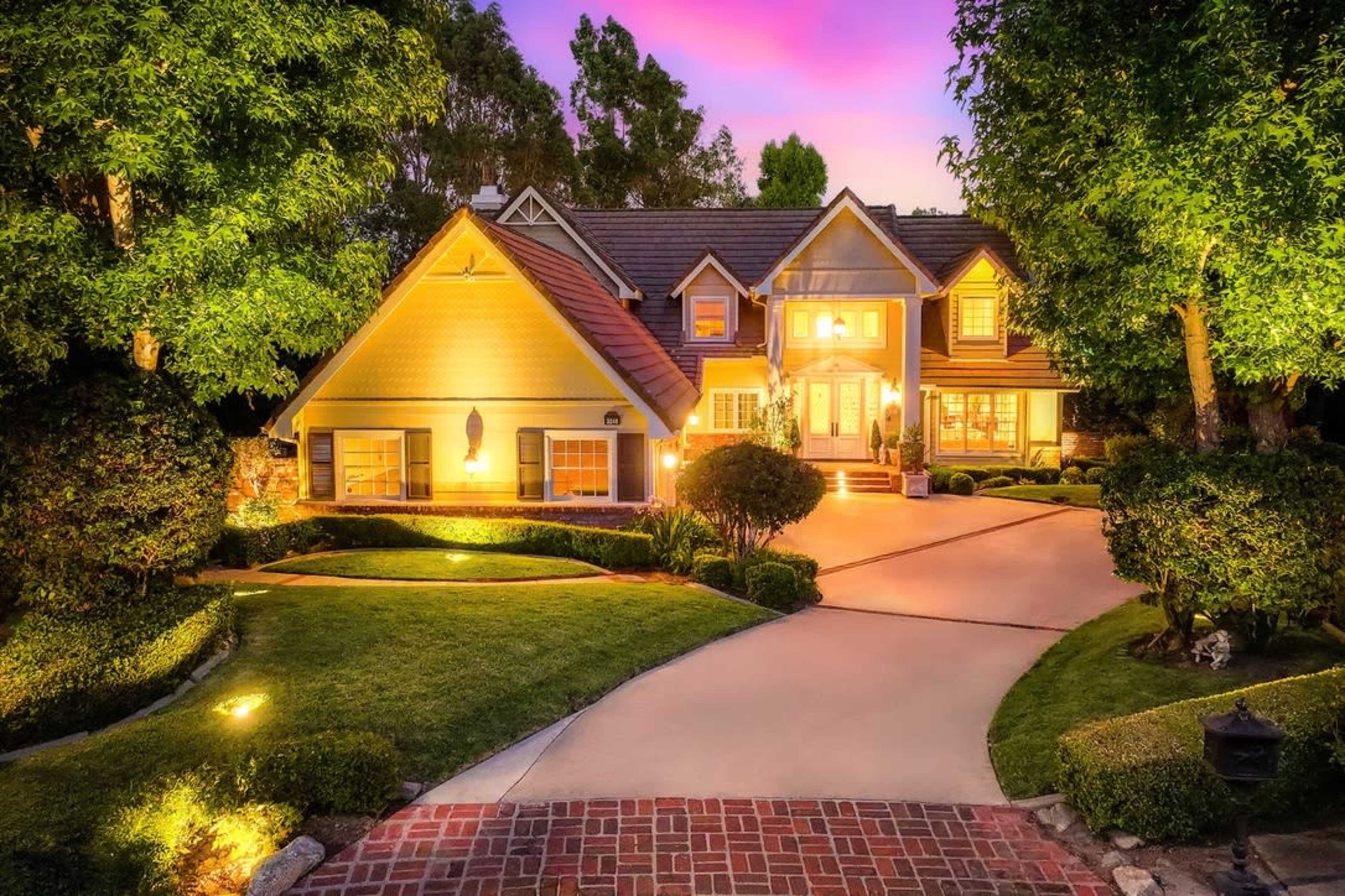 A large, well-lit house with a landscaped yard sits at the end of a paved driveway, surrounded by trees and shrubs under a colorful sunset sky.