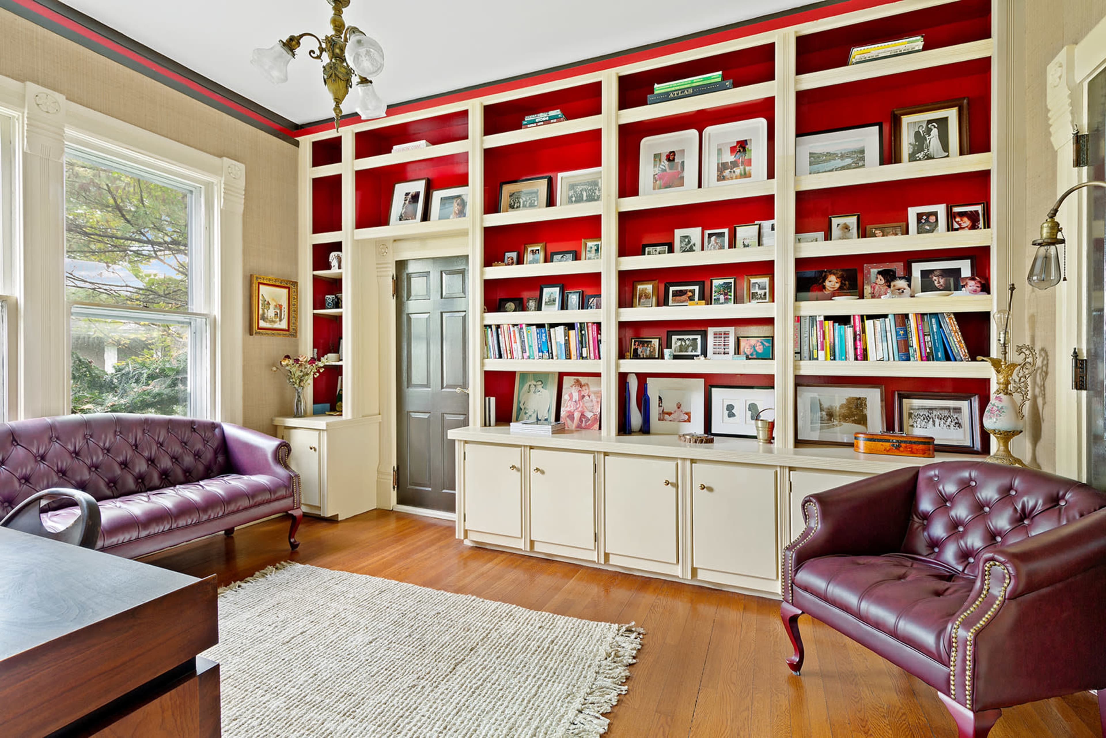 The image shows a room featuring two tufted chairs, a bookcase filled with books and framed photos, and a large window.
