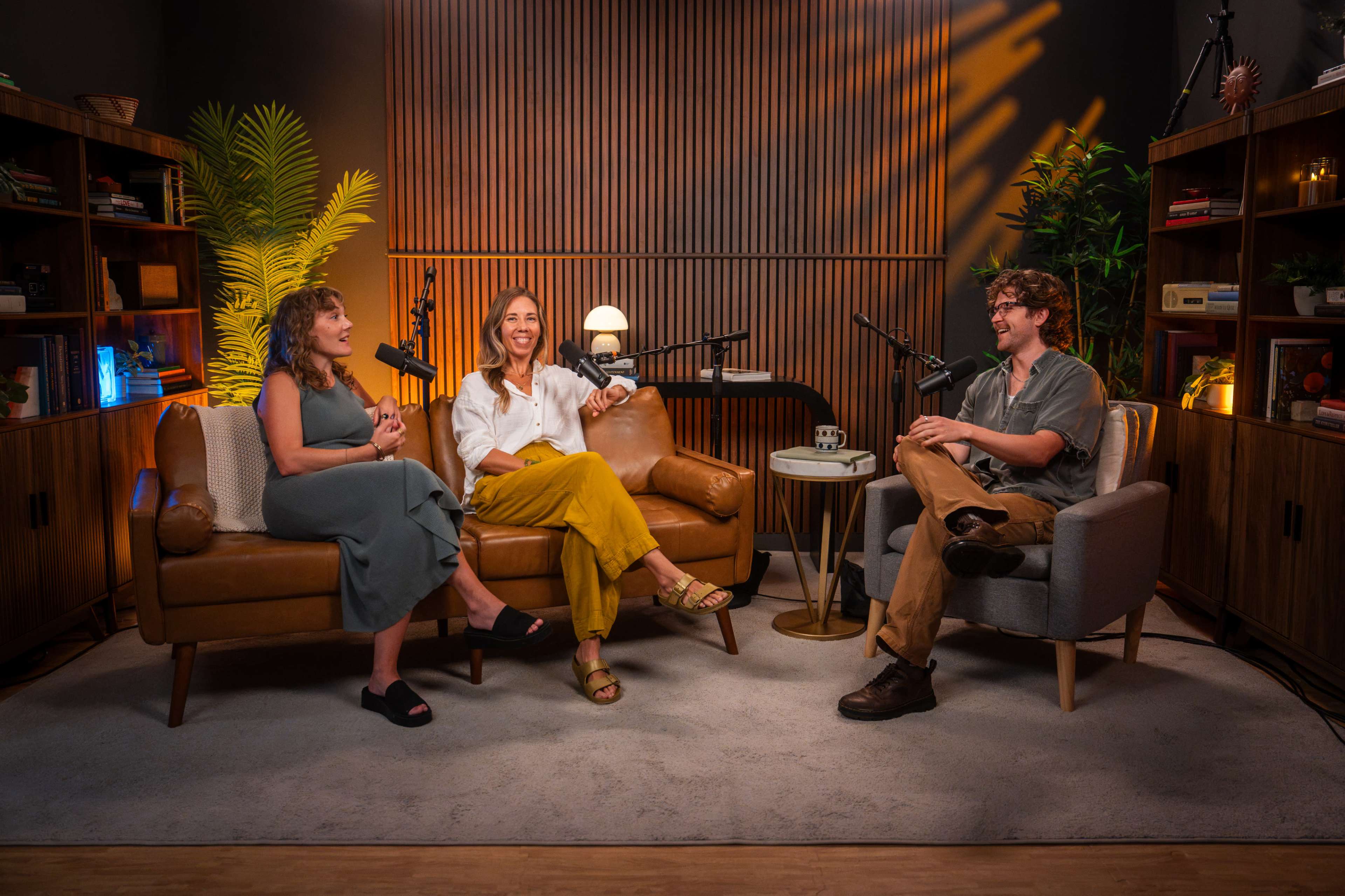 Three people are seated in a modern, well-lit studio with microphones in front of them, surrounded by bookshelves and plants.