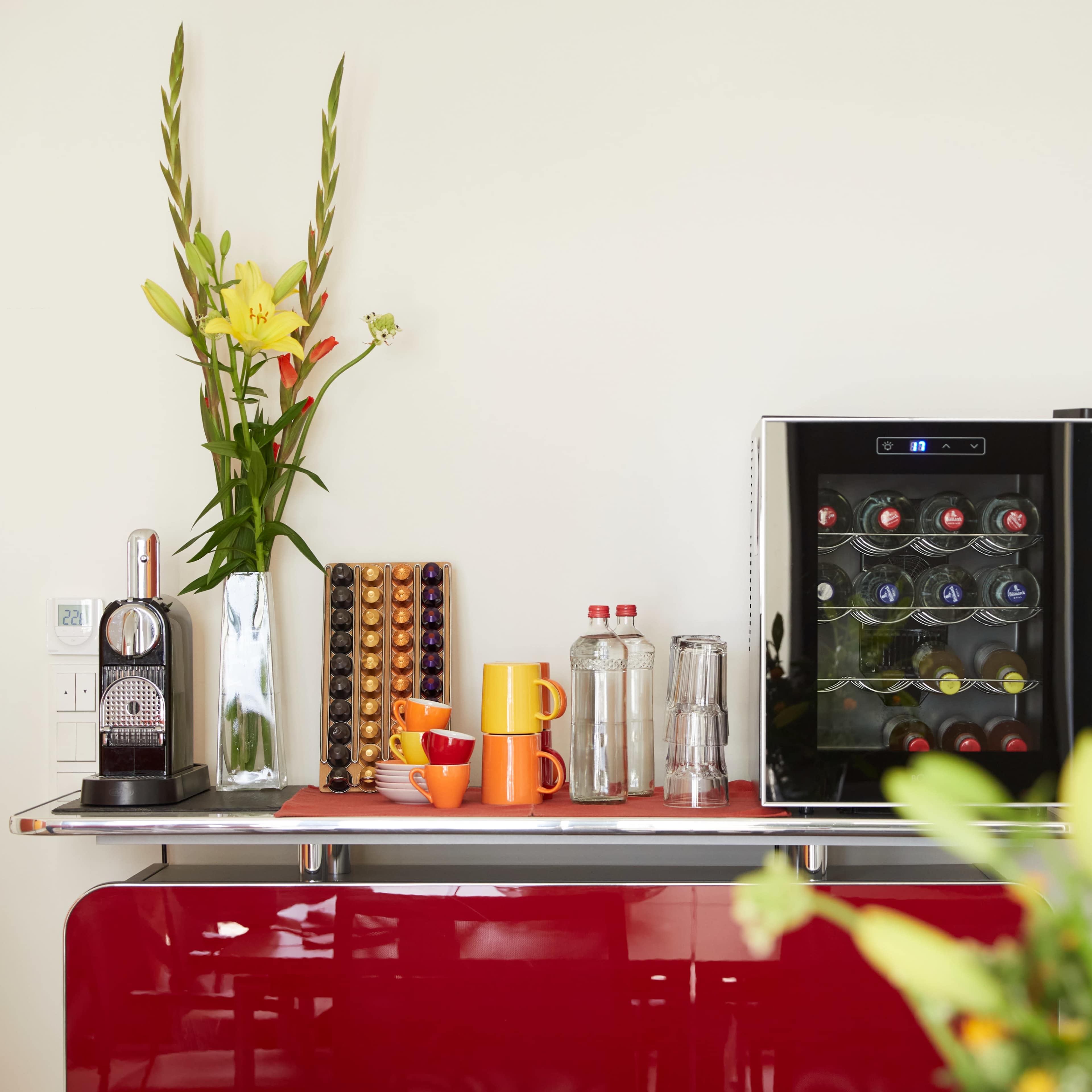 A bright kitchen counter displays a coffee machine, colorful mugs, and a wine cooler, accompanied by a vase of flowers.