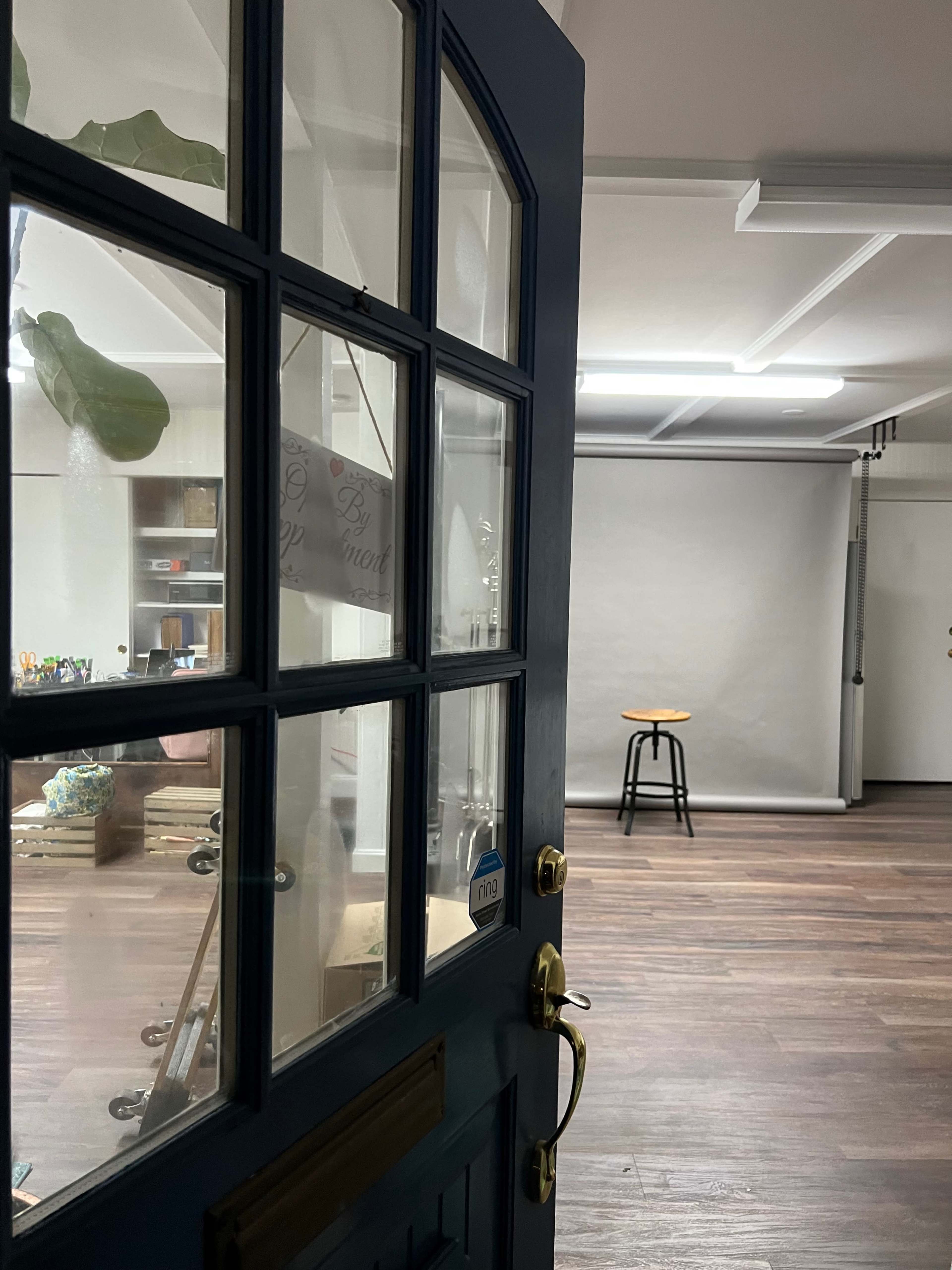 The image shows a partially open blue door leading into a sparsely furnished studio with a stool and a plain backdrop.