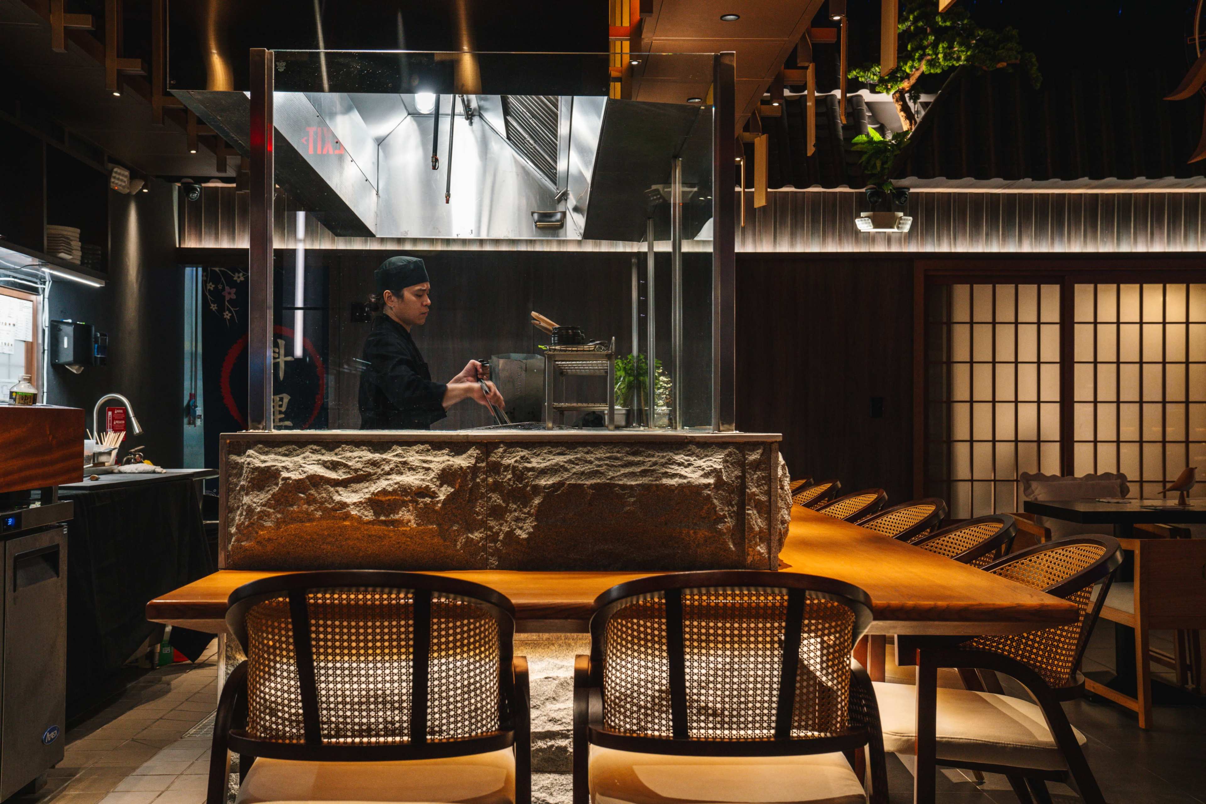 A chef stands behind a counter in a modern restaurant with a stone structure, surrounded by a series of empty wooden chairs.