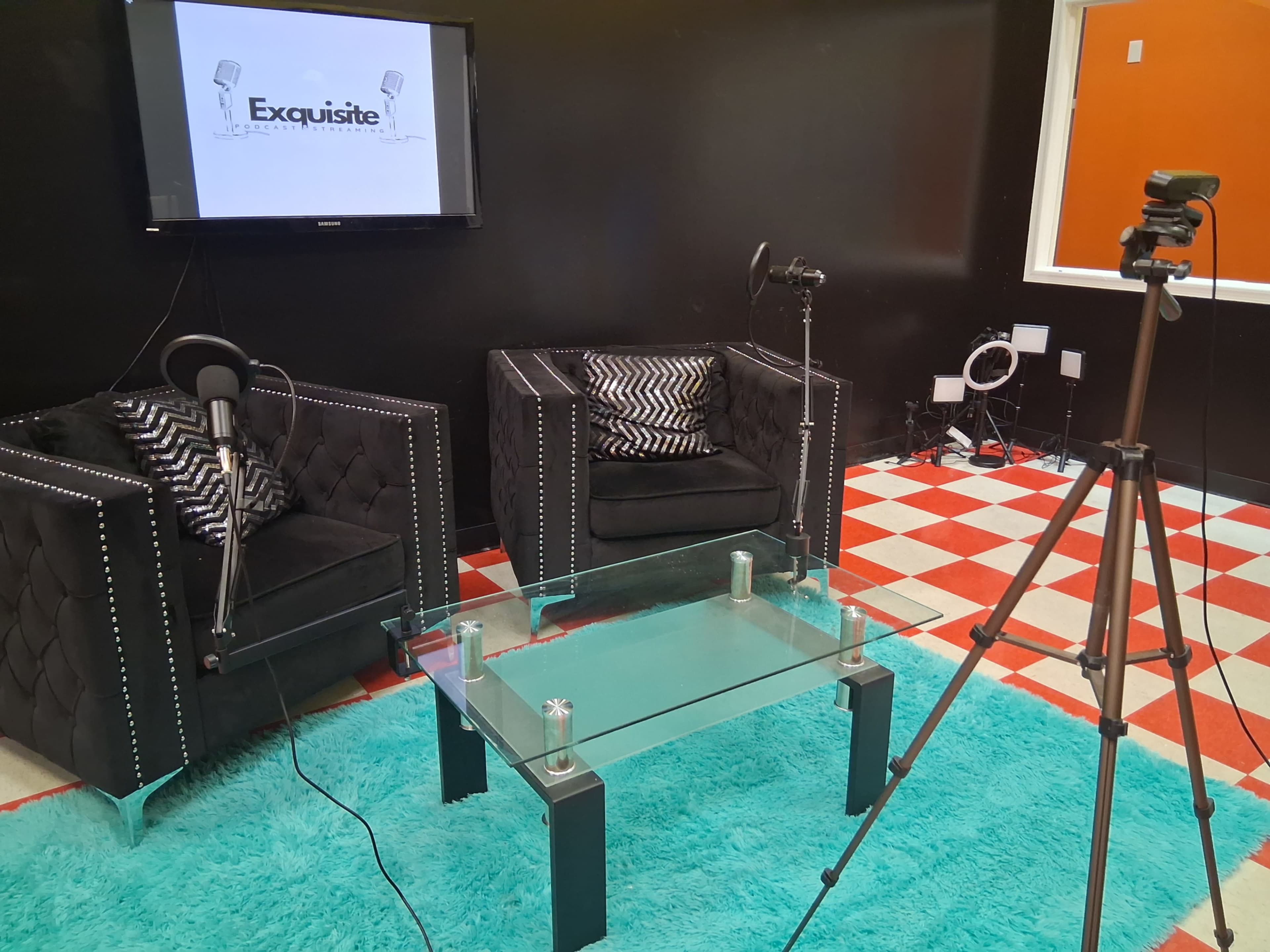 The image shows a modern interview setup featuring two black velvet chairs, a glass coffee table, a microphone on each chair, and a tripod with a camera, all against a black wall.