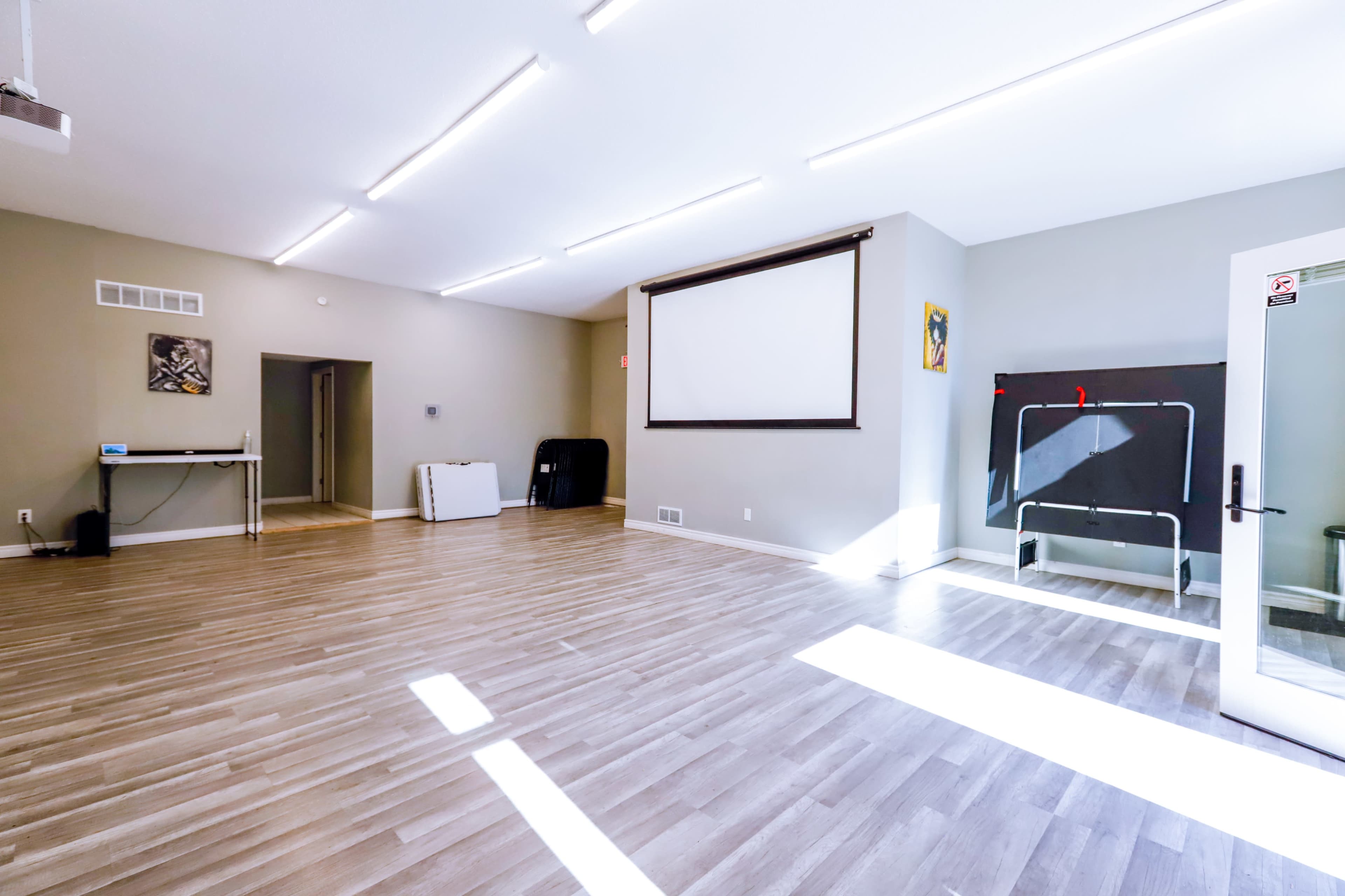 An empty, well-lit room with wooden flooring and a large projection screen on one wall.