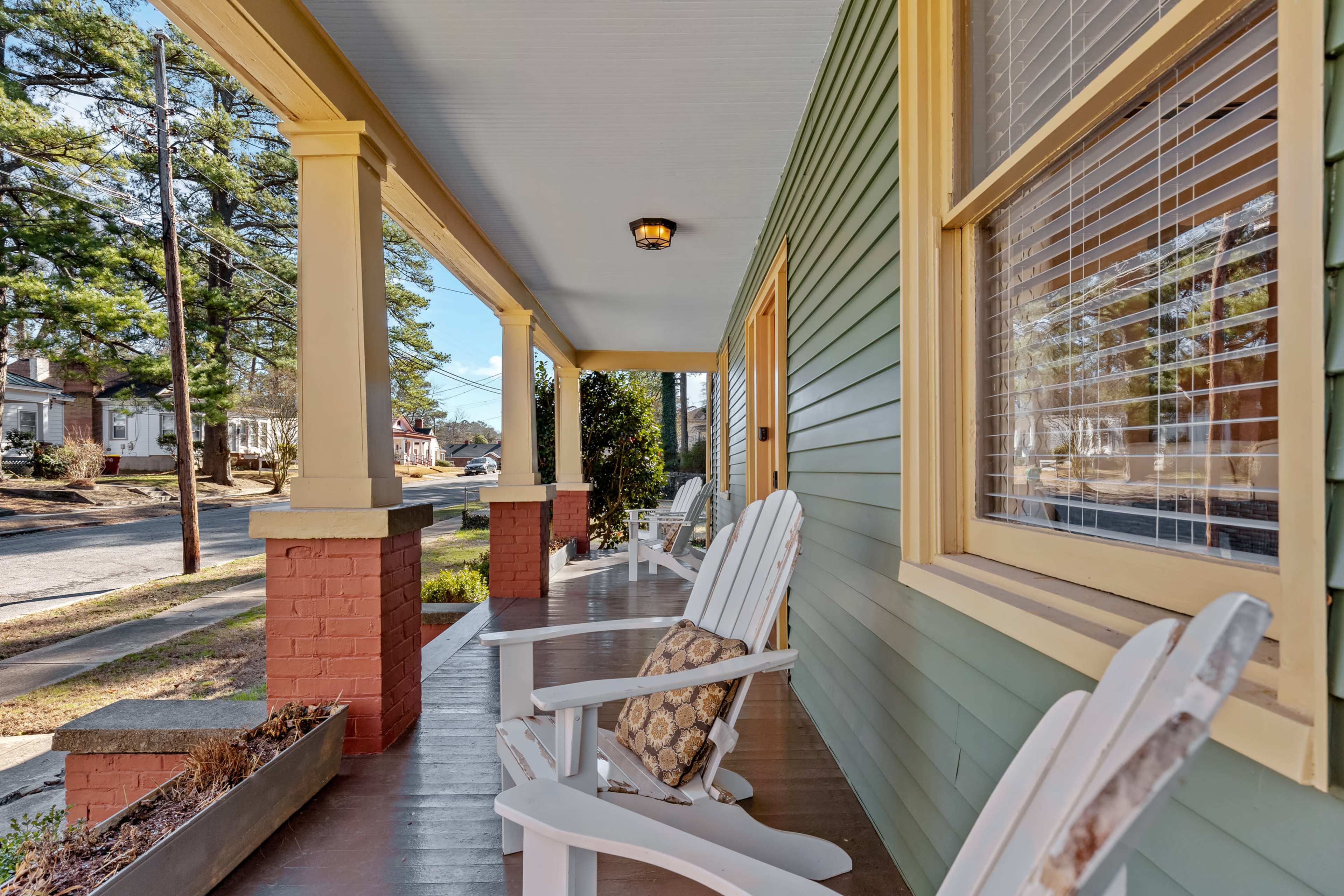 A porch with white rocking chairs and a decorative pillow, surrounded by green walls and yellow columns, overlooking a quiet street.