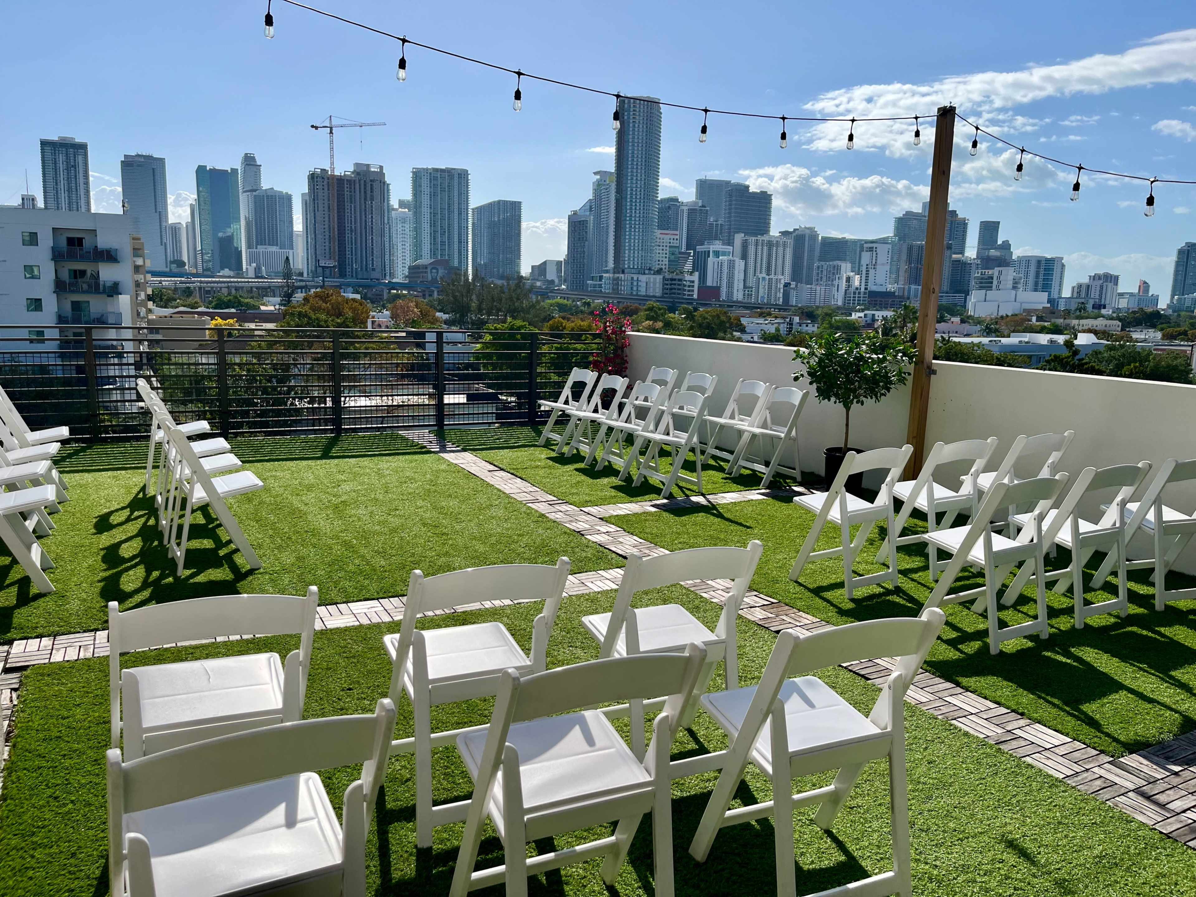 The scene shows a rooftop venue in an urban area, configured with rows of white chairs facing a skyline of tall buildings under a clear blue sky.