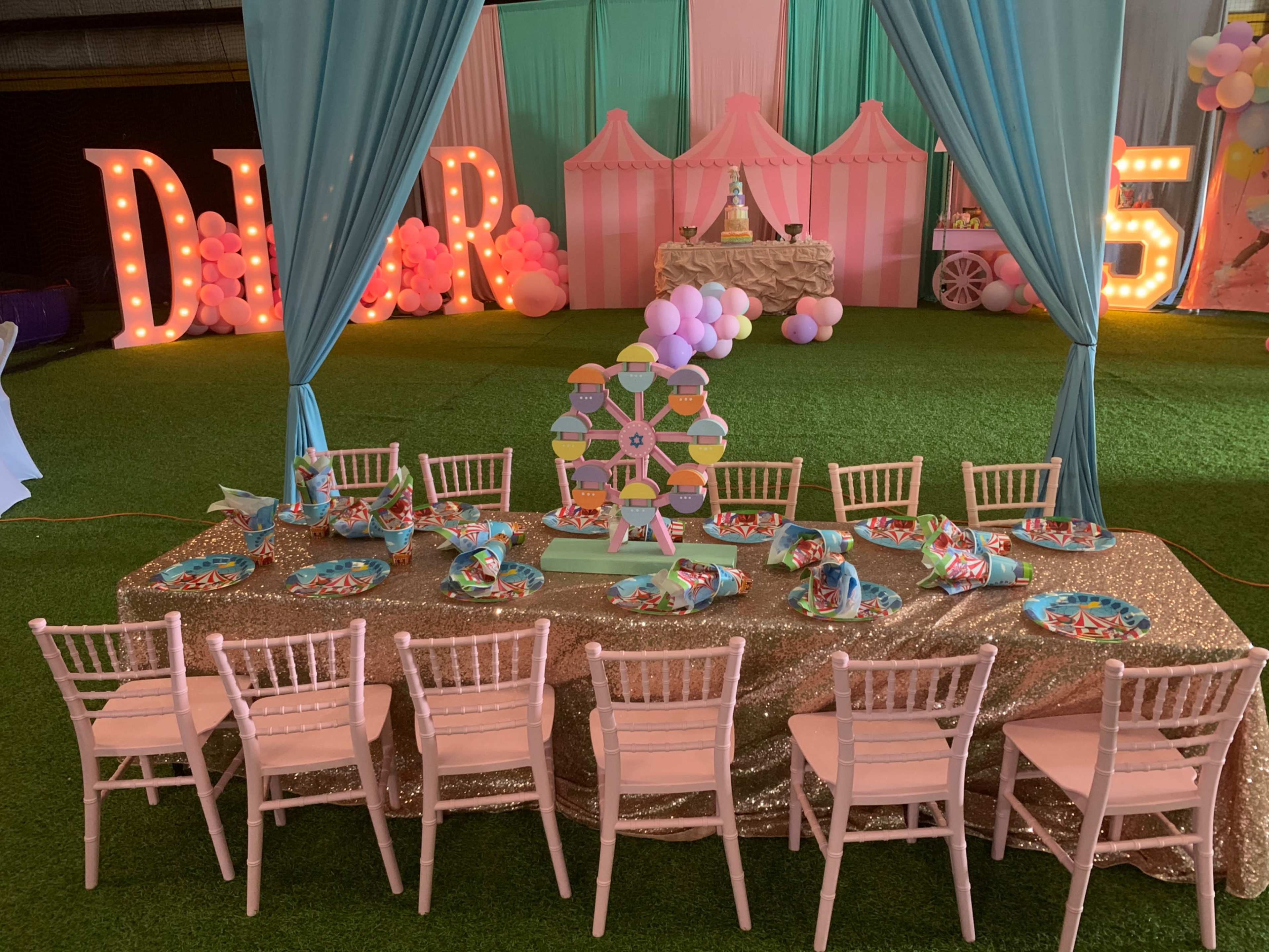 A decorated event space features a long table set with colorful party plates and chairs, surrounded by pastel drapes and festive decorations.