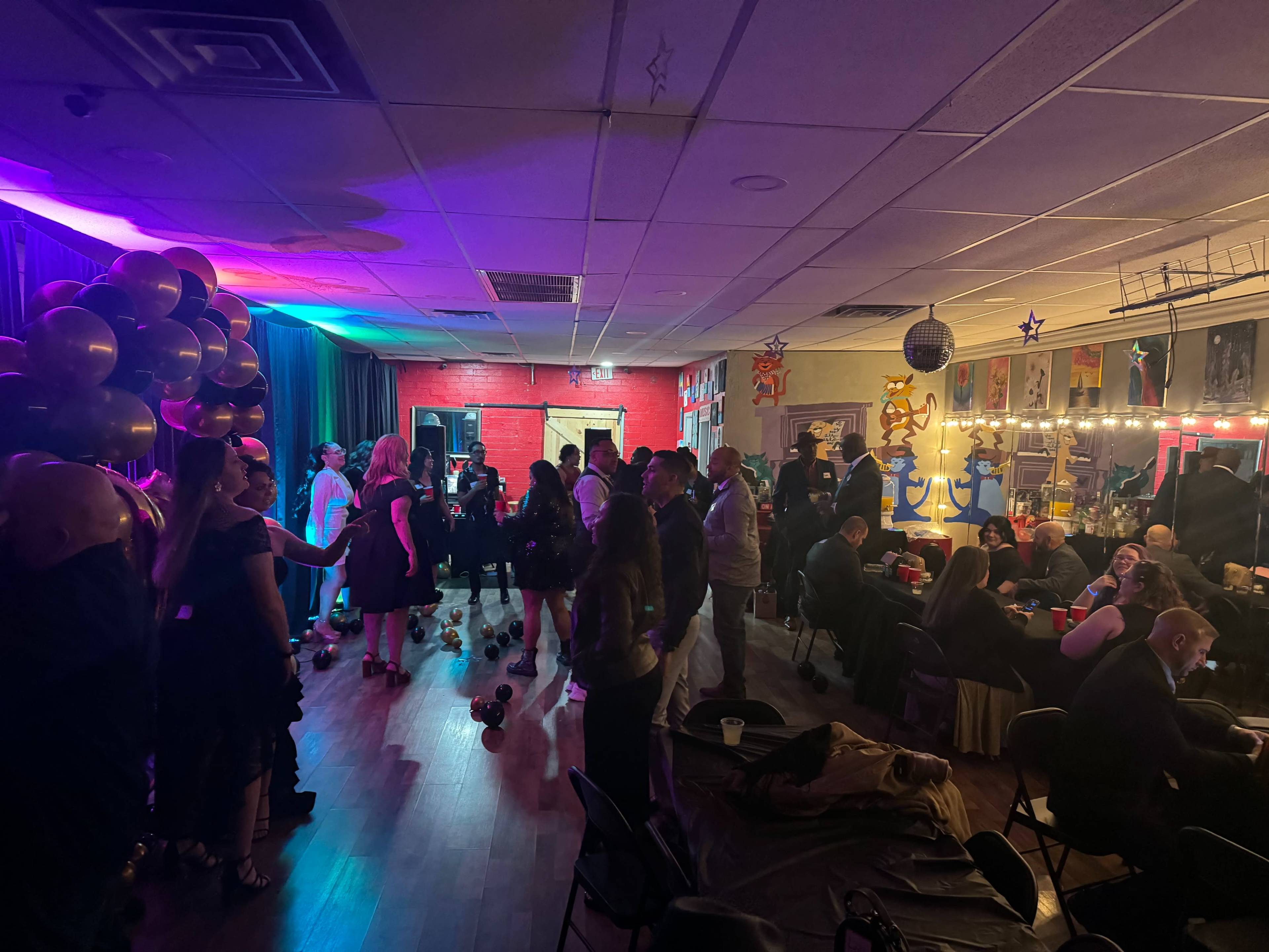 A lively event is taking place in a decorated room filled with people socializing, dancing, and standing around tables.