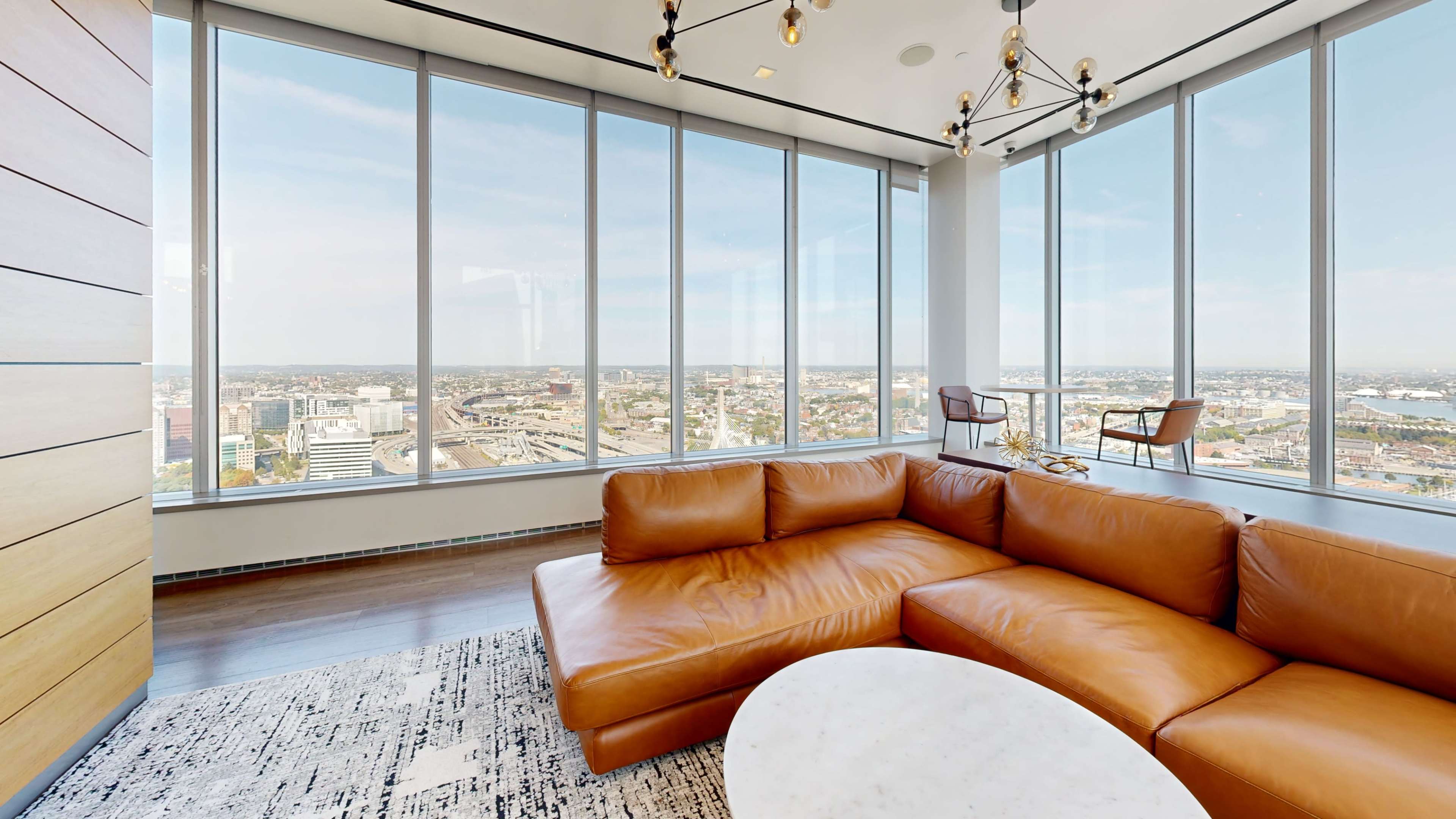The image shows a modern living space featuring a brown leather sectional sofa, a round marble coffee table, and large windows offering a panoramic view of a cityscape.