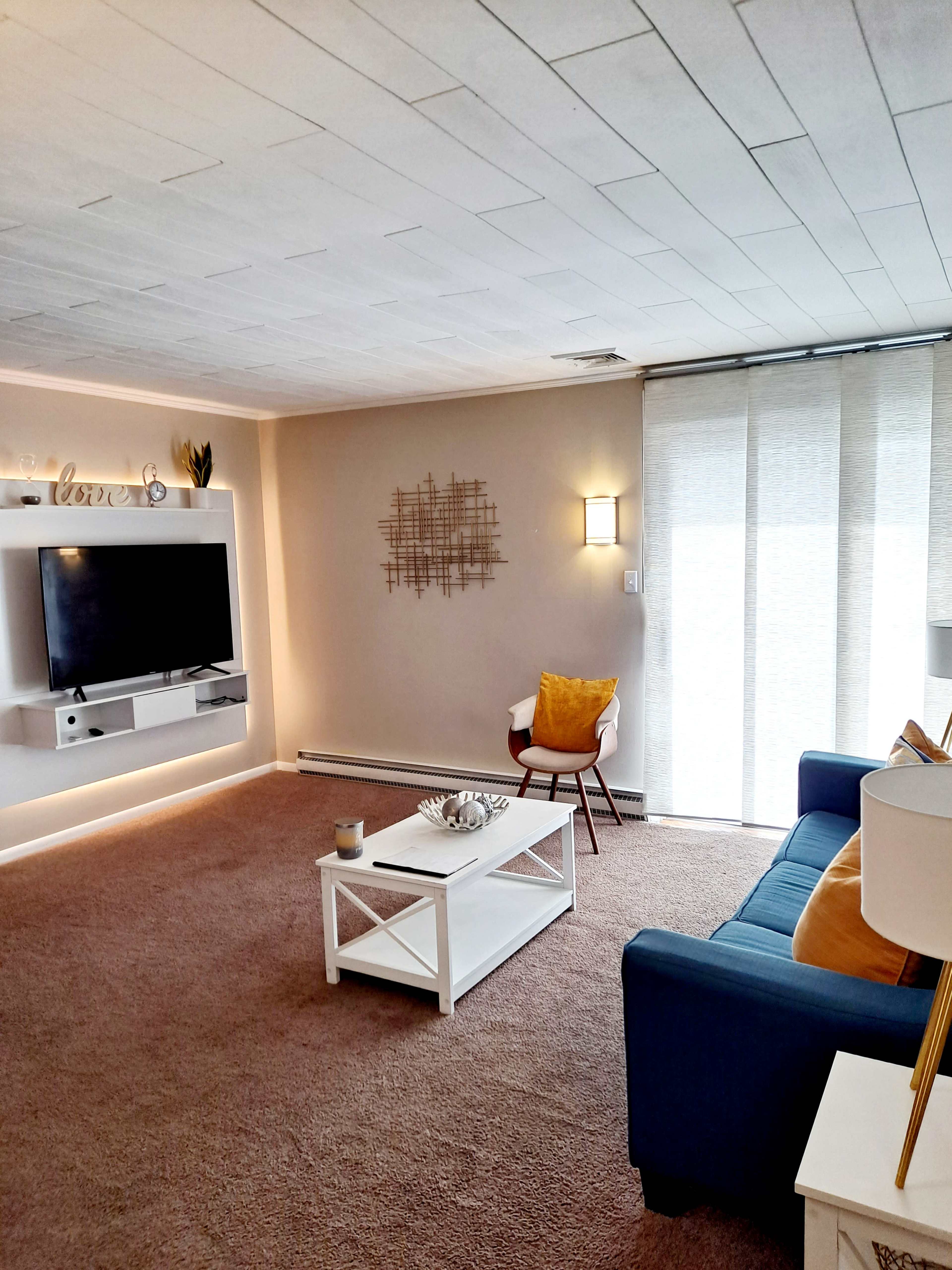 The image shows a cozy living room with a blue sofa, a white coffee table, a television mounted on the wall, and large windows covered with light, sheer curtains.