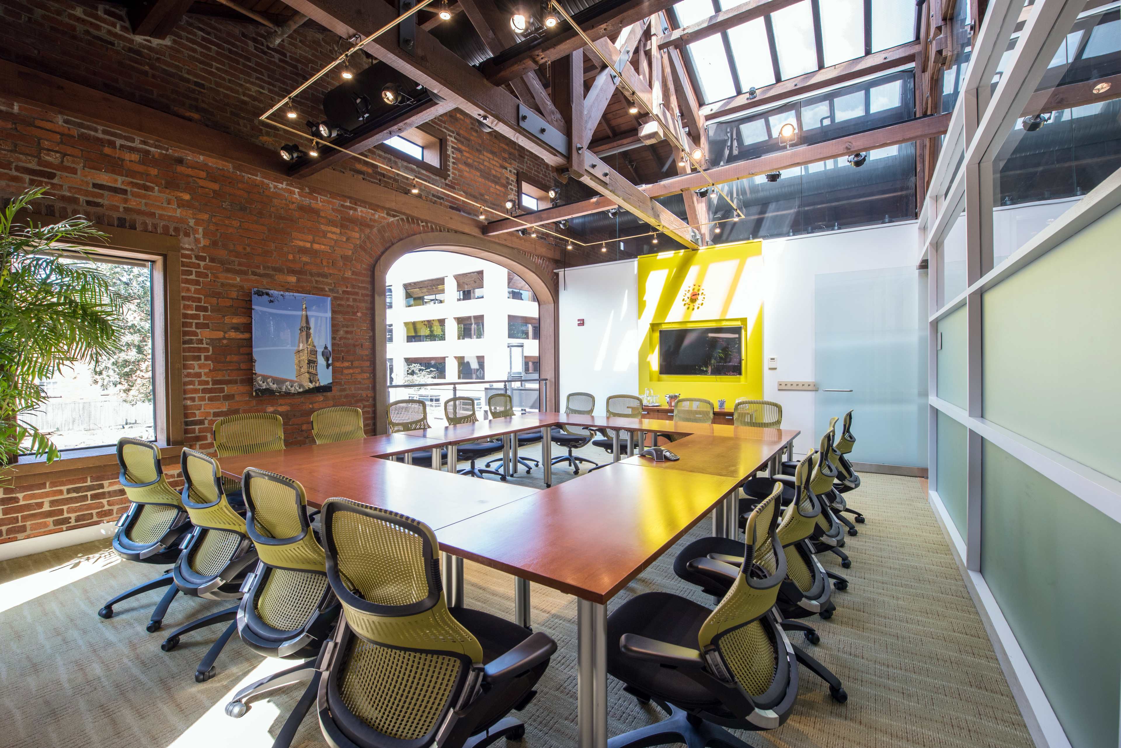 A spacious conference room features a long wooden table surrounded by ergonomic chairs, with large windows providing natural light and a view of a cityscape.