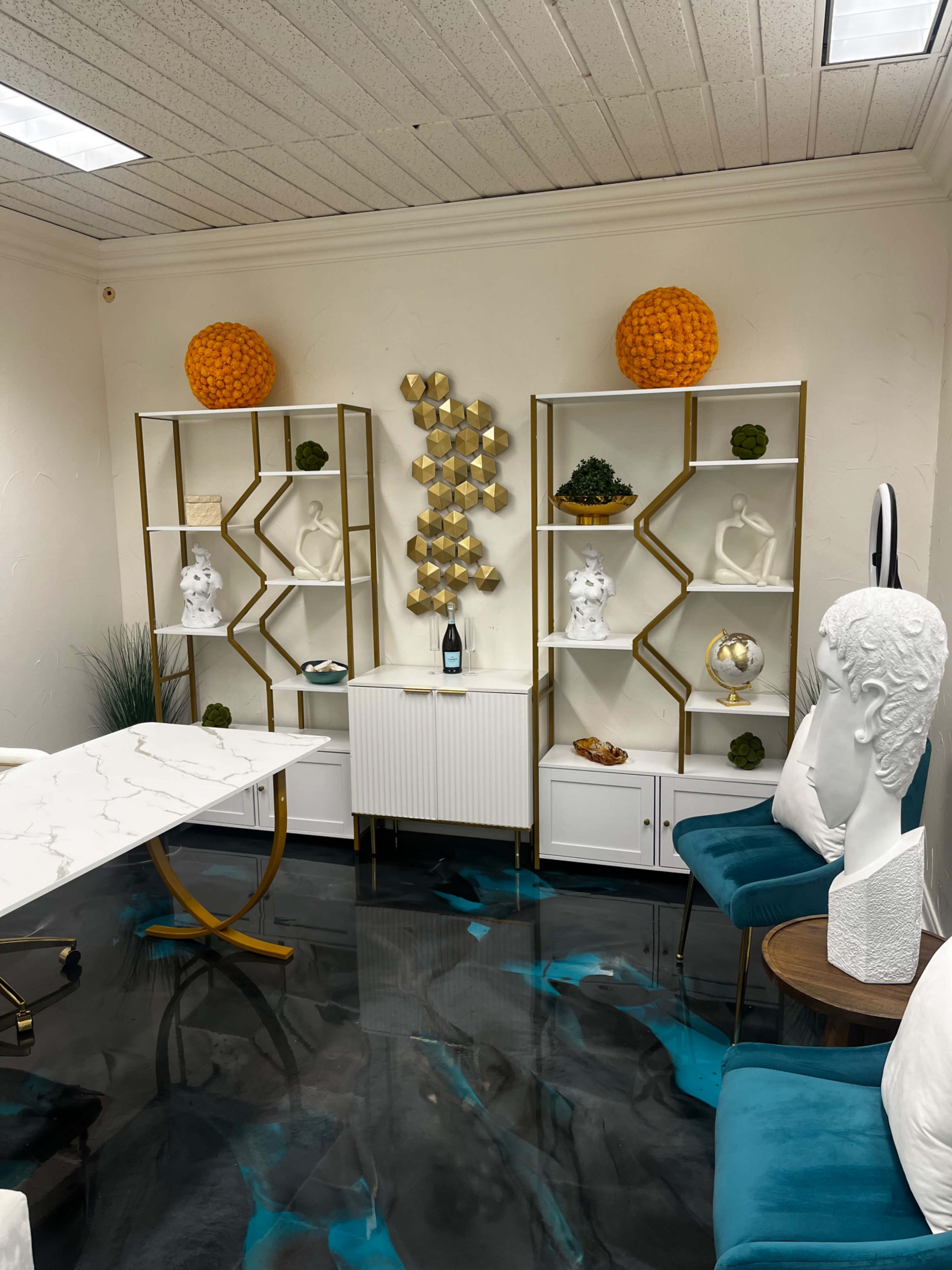 The image shows a modern office space with white and gold shelves displaying decorative items, a marble-topped desk, and teal upholstered chairs on a dark blue floor.