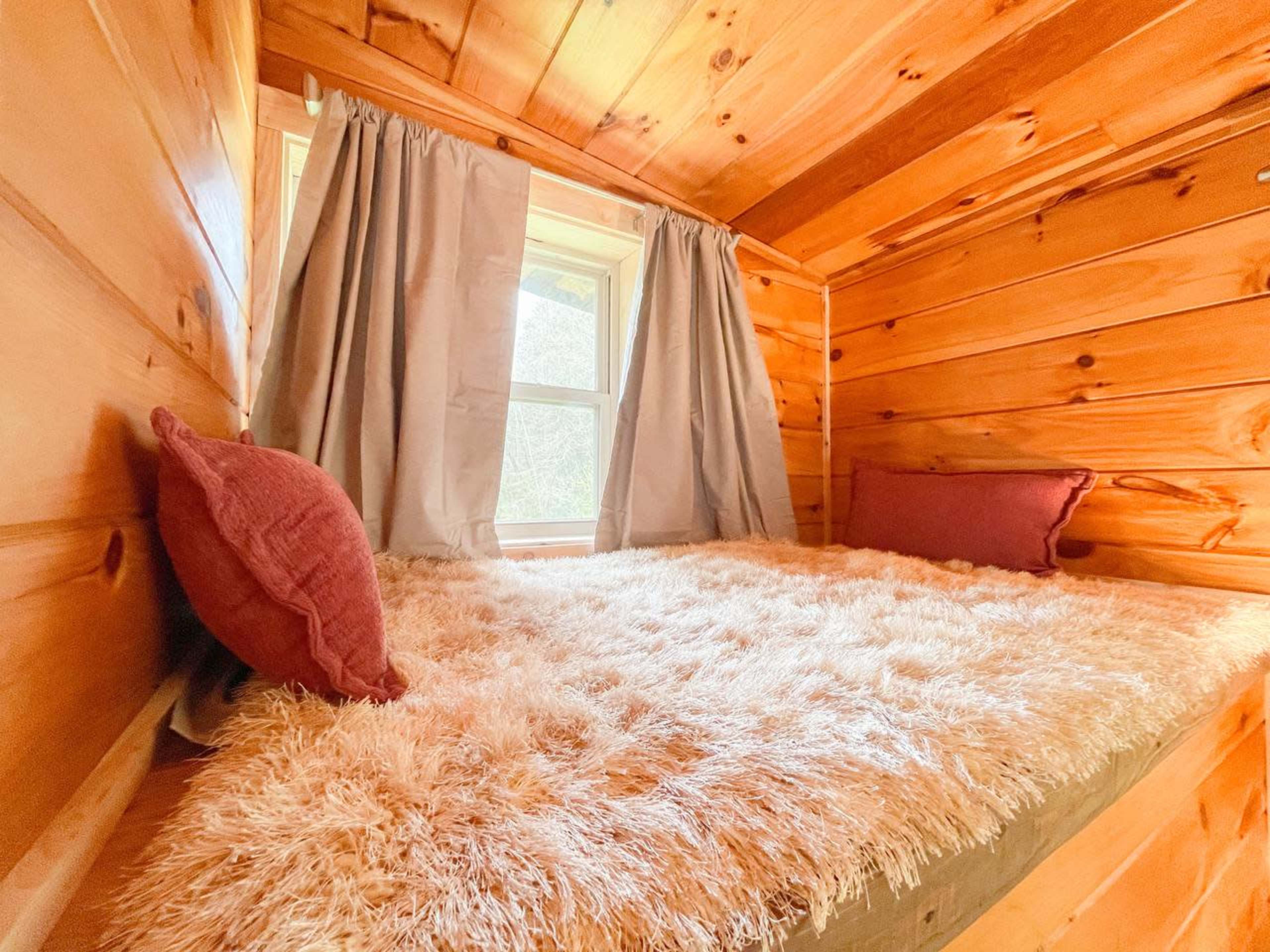 A cozy nook with a plush, shaggy bed covered in light-colored fabric, situated beneath a window with gray curtains in a wooden interior.