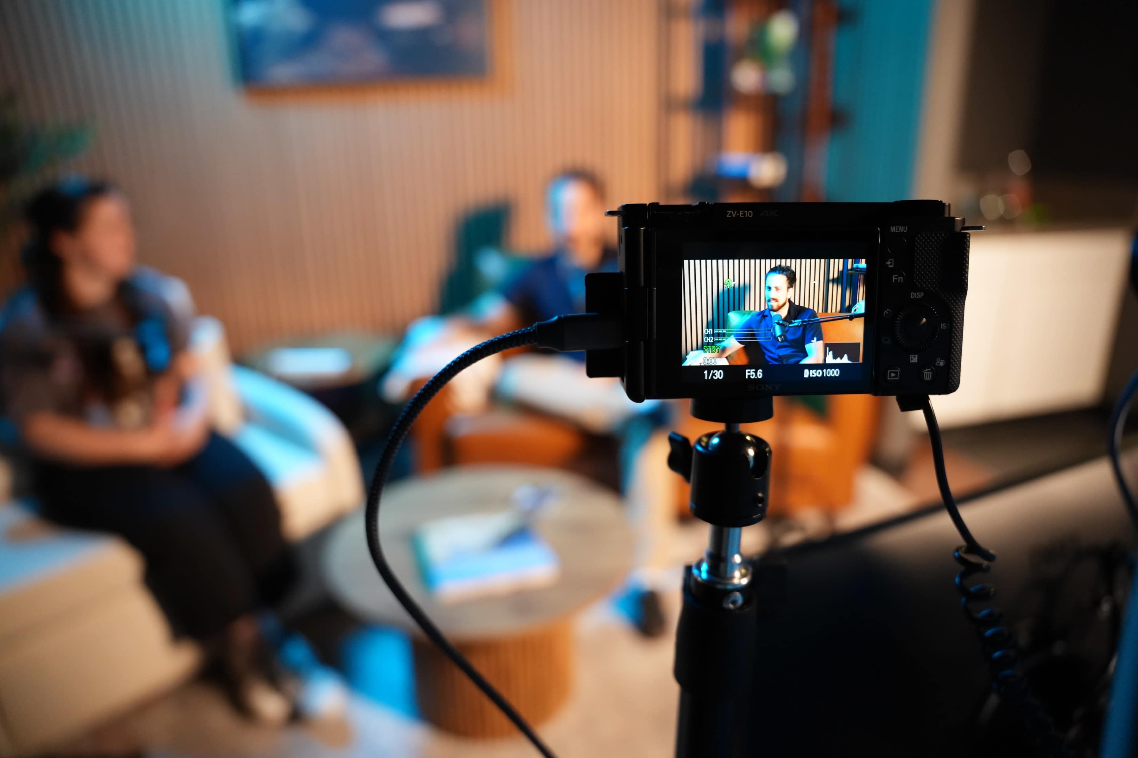 A camera on a tripod captures two people seated on a sofa in a well-lit interior space.