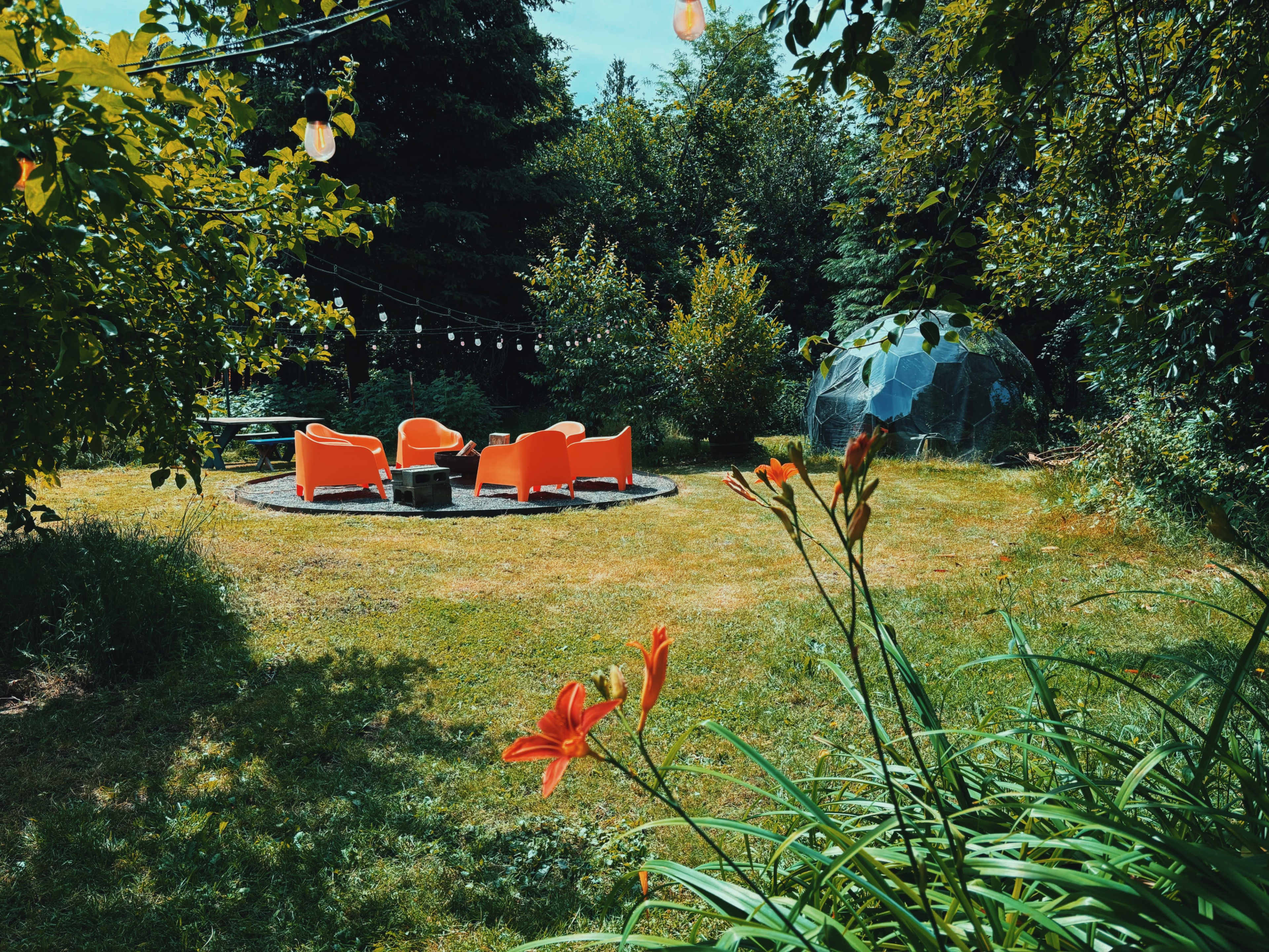 A circular seating area with four orange chairs surrounds a fire pit, set in a grassy garden with trees and a geodesic dome in the background.