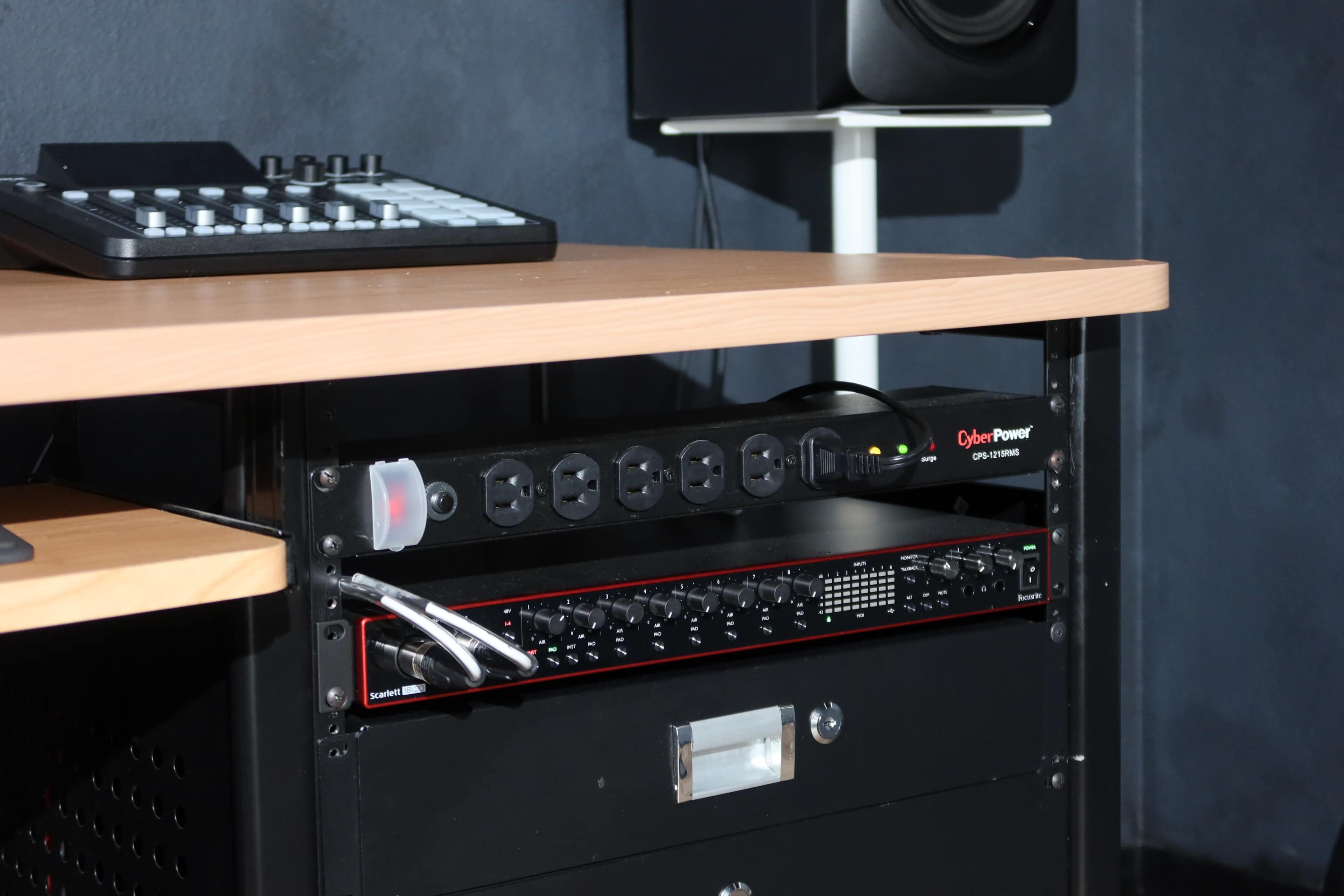 The image shows a black audio rack with power distribution, a mixer, and recording equipment on a wooden desk in a home studio.