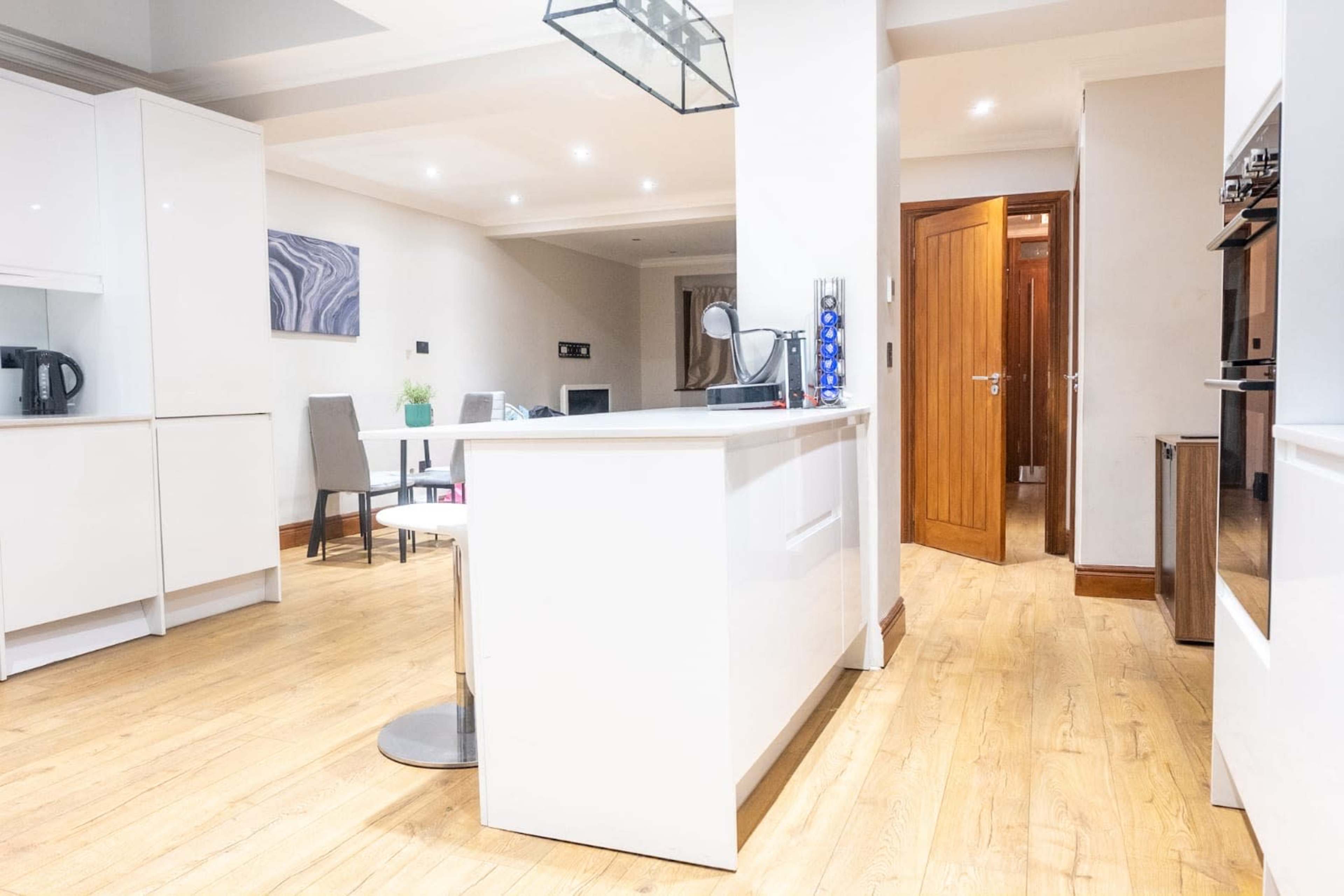 The image shows a modern kitchen and dining area featuring white cabinetry, a wooden floor, and a doorway leading to another room.