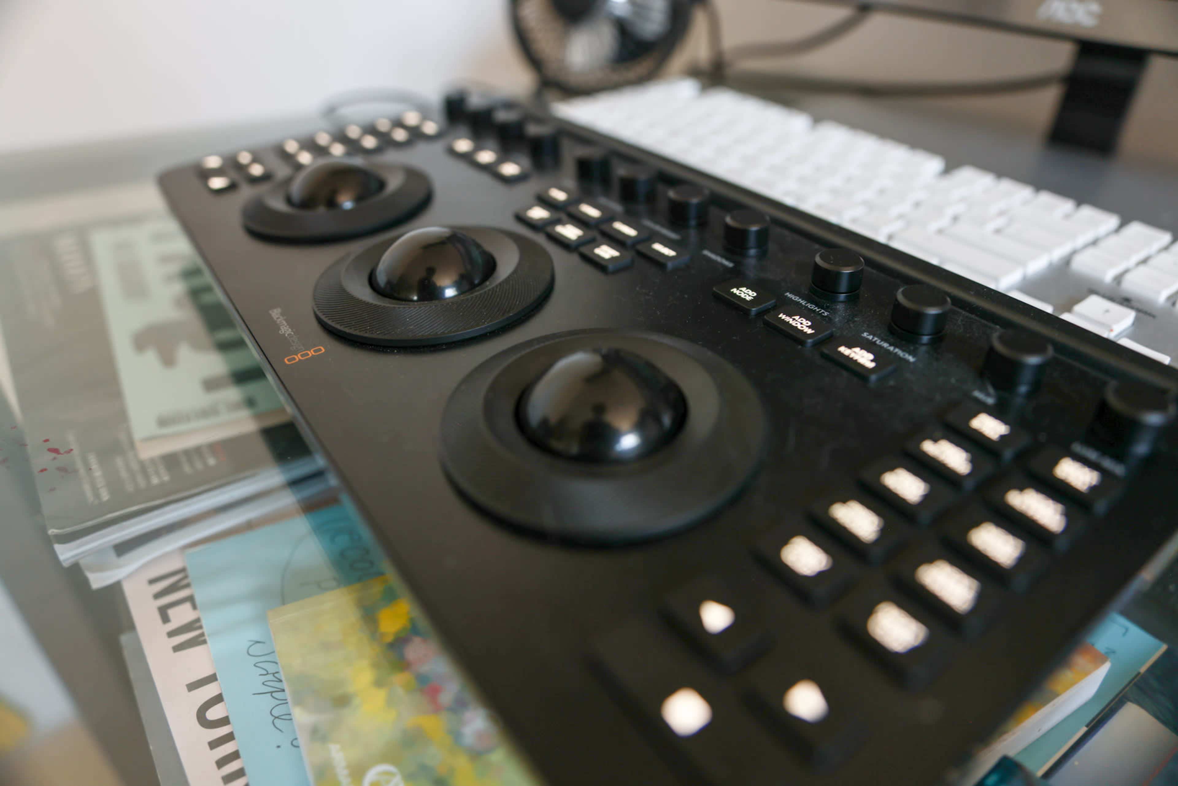 The image shows a close-up of a music production controller featuring multiple knobs and buttons on a desk with a computer keyboard in the background.