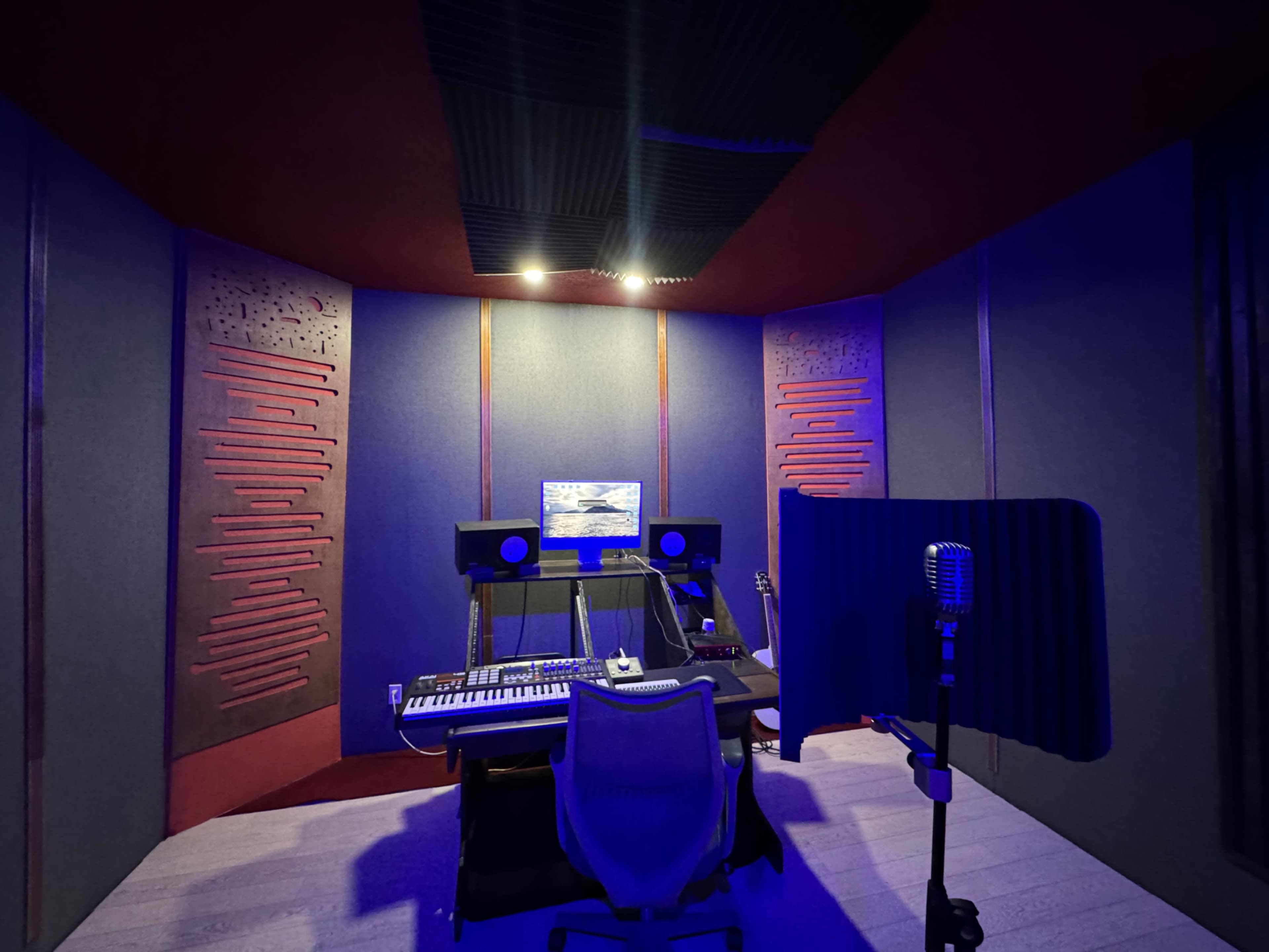 The image shows a music recording studio with acoustic panels, a desk with audio equipment, and a microphone setup in a corner.