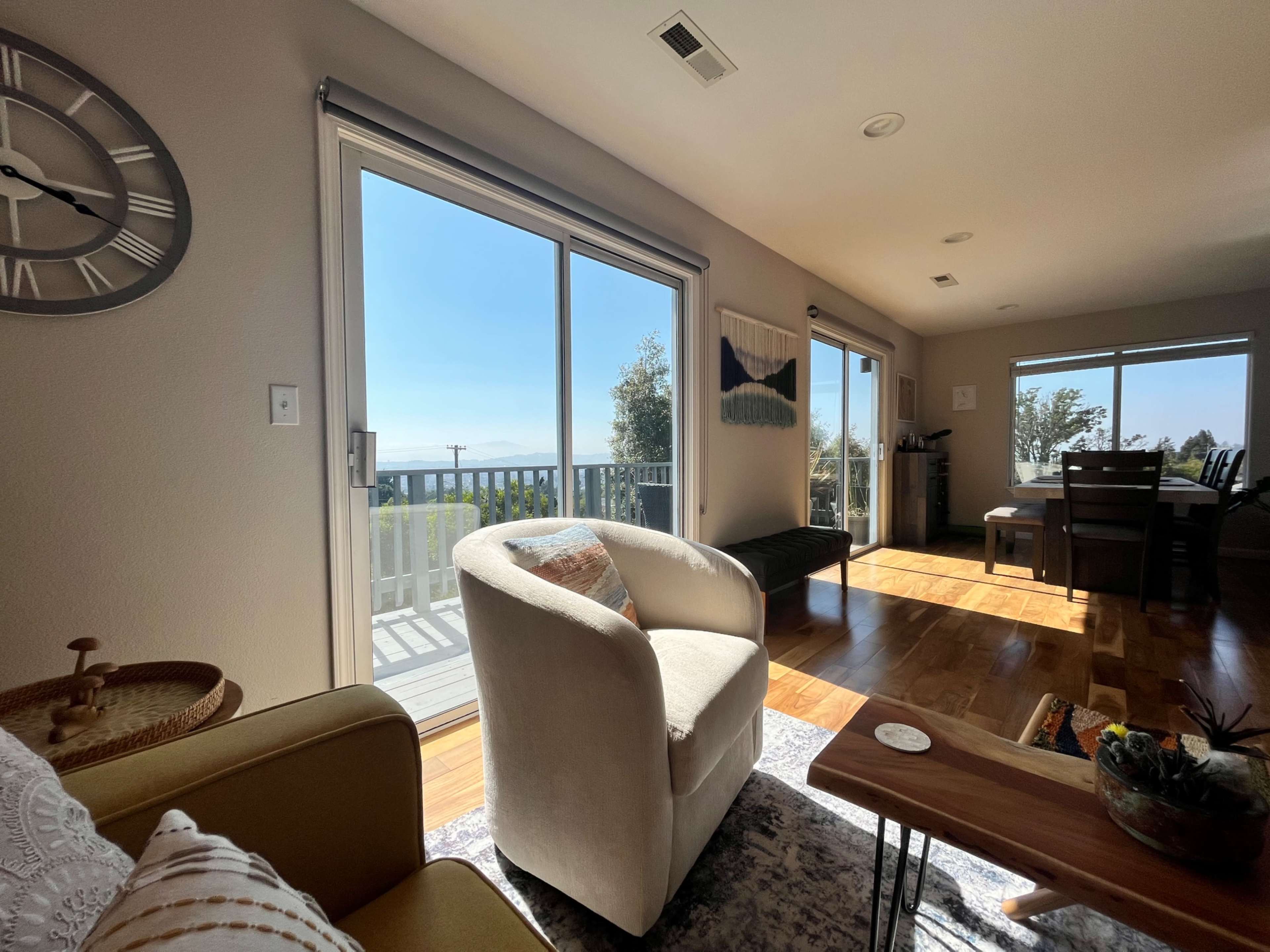 A bright living room with large windows overlooking a scenic view, featuring a white armchair, a small wooden table, and a dining area in the background.