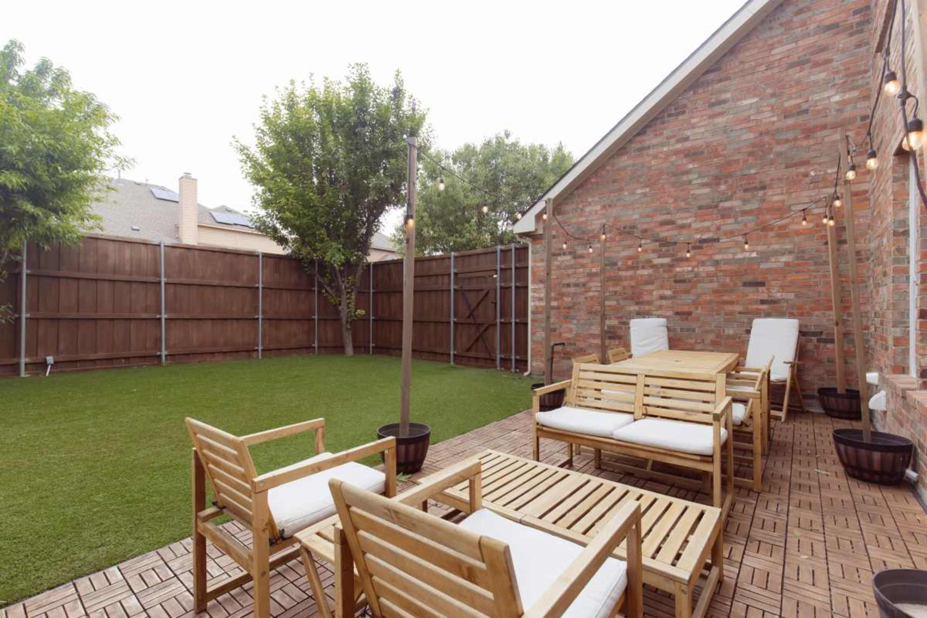 The image shows a backyard patio area with wooden furniture, including chairs and a table, surrounded by a lawn and a wooden fence.