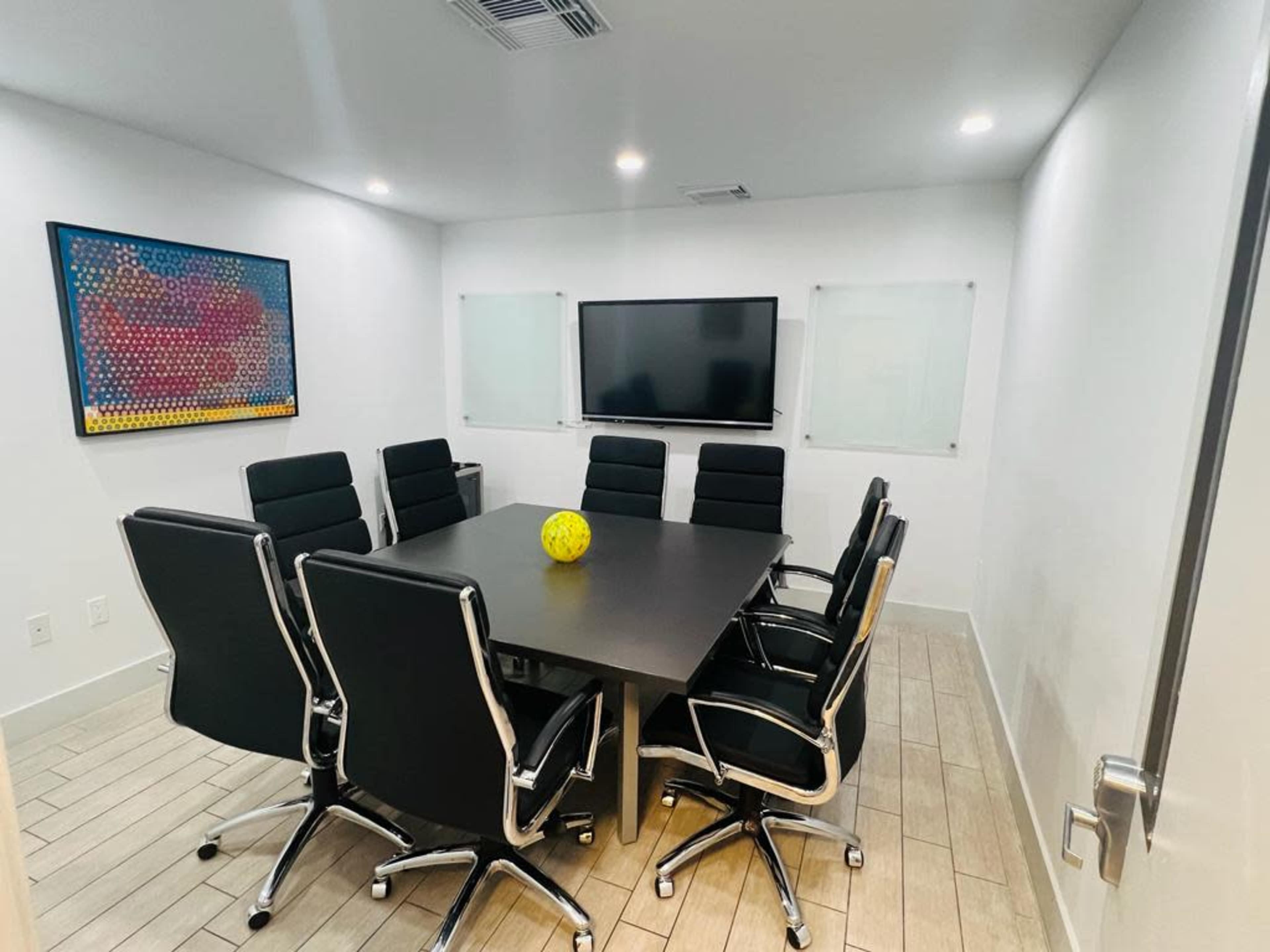A modern conference room features a large black table surrounded by eight black chairs and a yellow decorative item at its center, with a television and whiteboards on the walls.