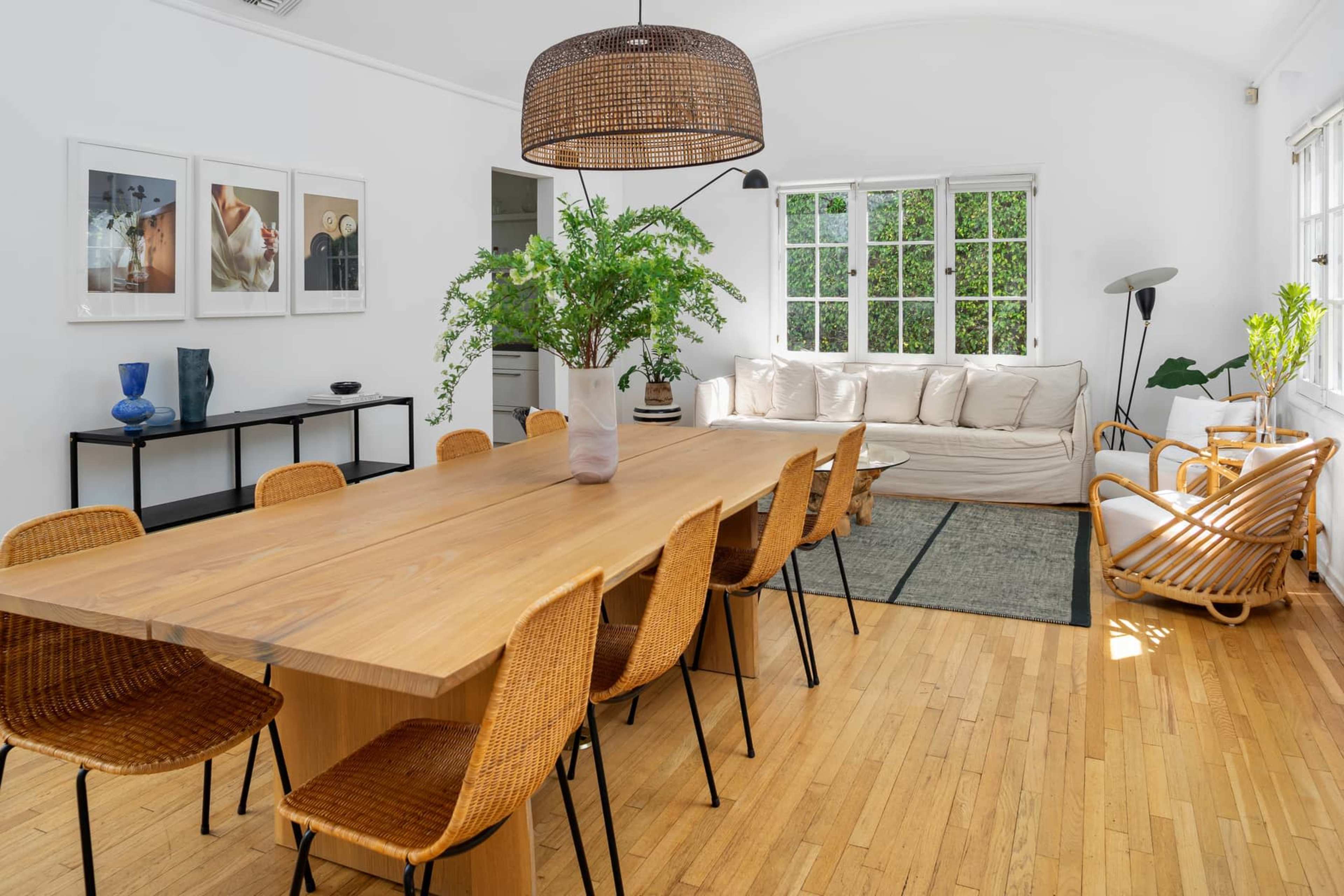A spacious dining area features a long wooden table surrounded by woven chairs, with a large pendant light overhead and a cozy seating area in the background.