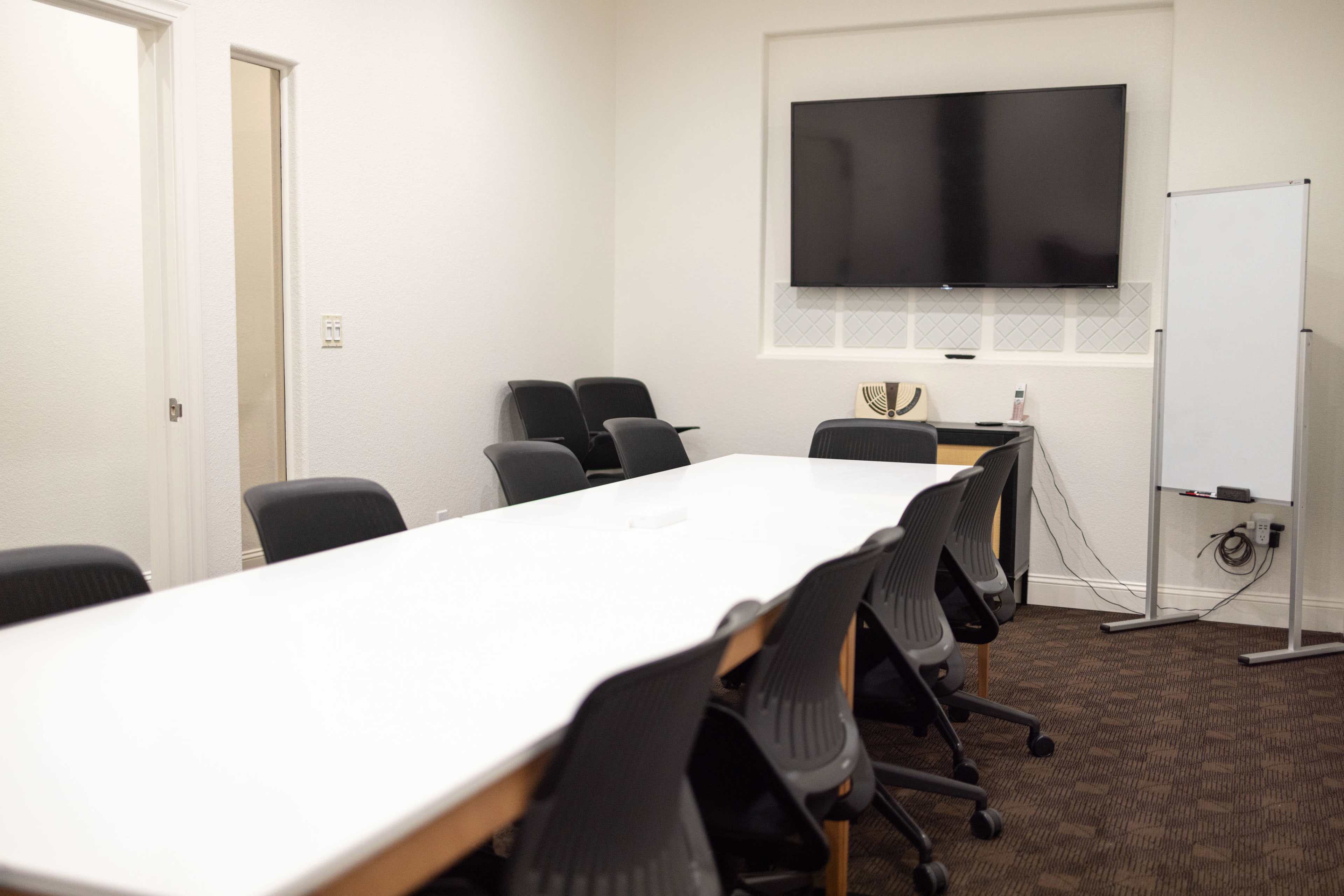 A conference room features a long white table surrounded by black chairs, with a large display screen mounted on the wall and a whiteboard nearby.