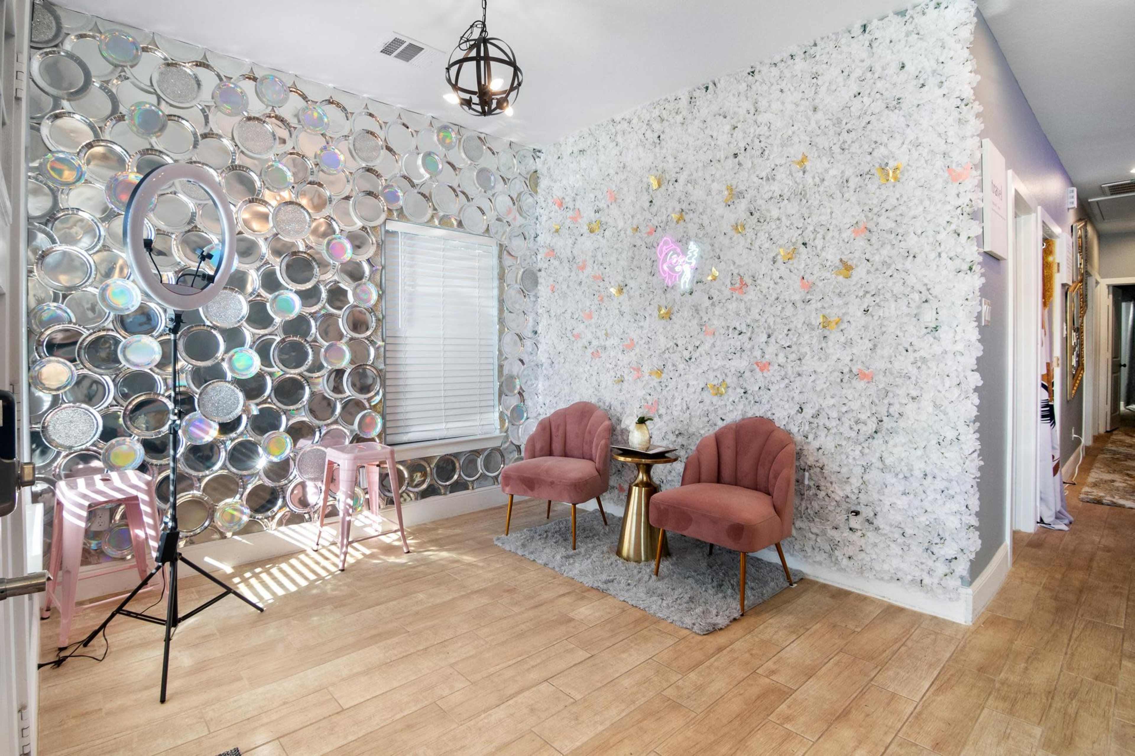 The scene features a well-lit interior with a mirrored wall on one side, a floral wallpaper on the other, and two pink chairs beside a small table.